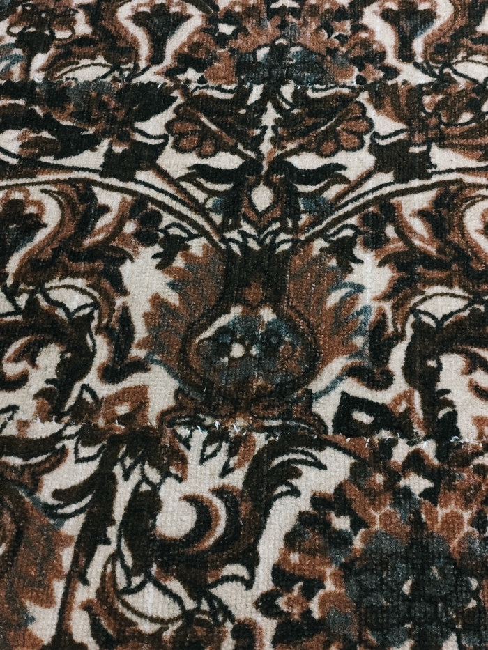 A close-up shot reveals a patterned carpet featuring brown, black, and beige tones. The design appears to be a floral or ornamental motif with repeating elements. Stitching is visible on the carpet, suggesting repairs or seams. The carpet's texture appears soft and plush. The overall impression is one of a well-worn, patterned textile.