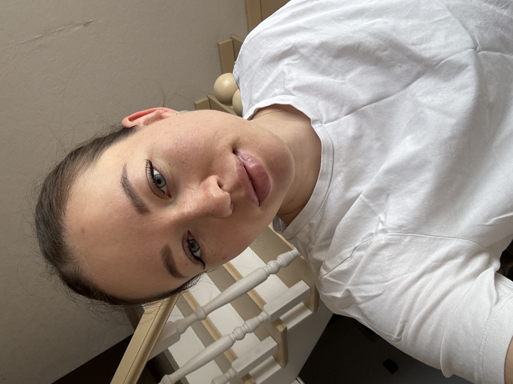 A young woman with fair skin, brown hair pulled back, and light-colored eyes is looking down at the camera. She is wearing a white t-shirt. In the background is a set of white stairs with wooden railings. It is difficult to verify whether this image was taken in Ферма, Russia.