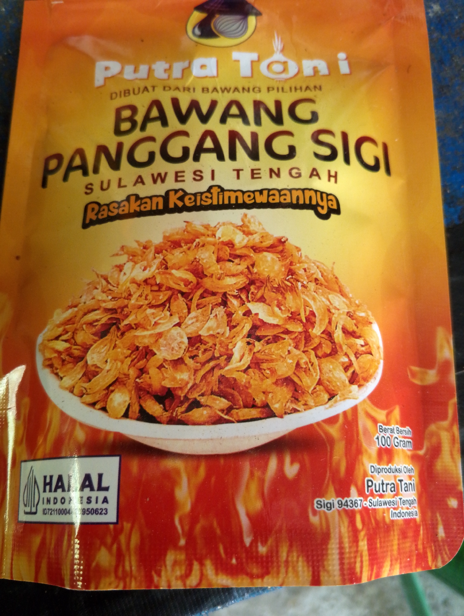 The image is a close-up of a package of fried onions. The packaging is primarily orange and yellow with flames depicted at the bottom. The brand name "Putra Toni" is prominently displayed in large white font at the top. Below this, it states "DIBUAT DARI BAWANG PILIHAN" (Made from selected onions). Further down, the product name "BAWANG PANGGANG SIGI" (Sigi Roasted Onions) is written in large, dark, textured font. Underneath that, "SULAWESI TENGAH" (Central Sulawesi) is written. A wavy text reads "Rasakan Keistimewaannya" (Taste its specialty). A white bowl filled with fried onions is pictured in the center of the package.
In the bottom left corner, a Halal certification is shown with the text "HALAL INDONESIA" and "ID721100044950623". In the bottom right, it states "Berat Bersih 100 Gram" (Net Weight 100 Grams), "Diproduksi Oleh Putra Tani" (Produced by Putra Tani), and "Sigi 94367-Sulawesi Tengah Indonesia".
The image does not provide any information or visual cues to determine the location within the city of Tomohon, Indonesia.