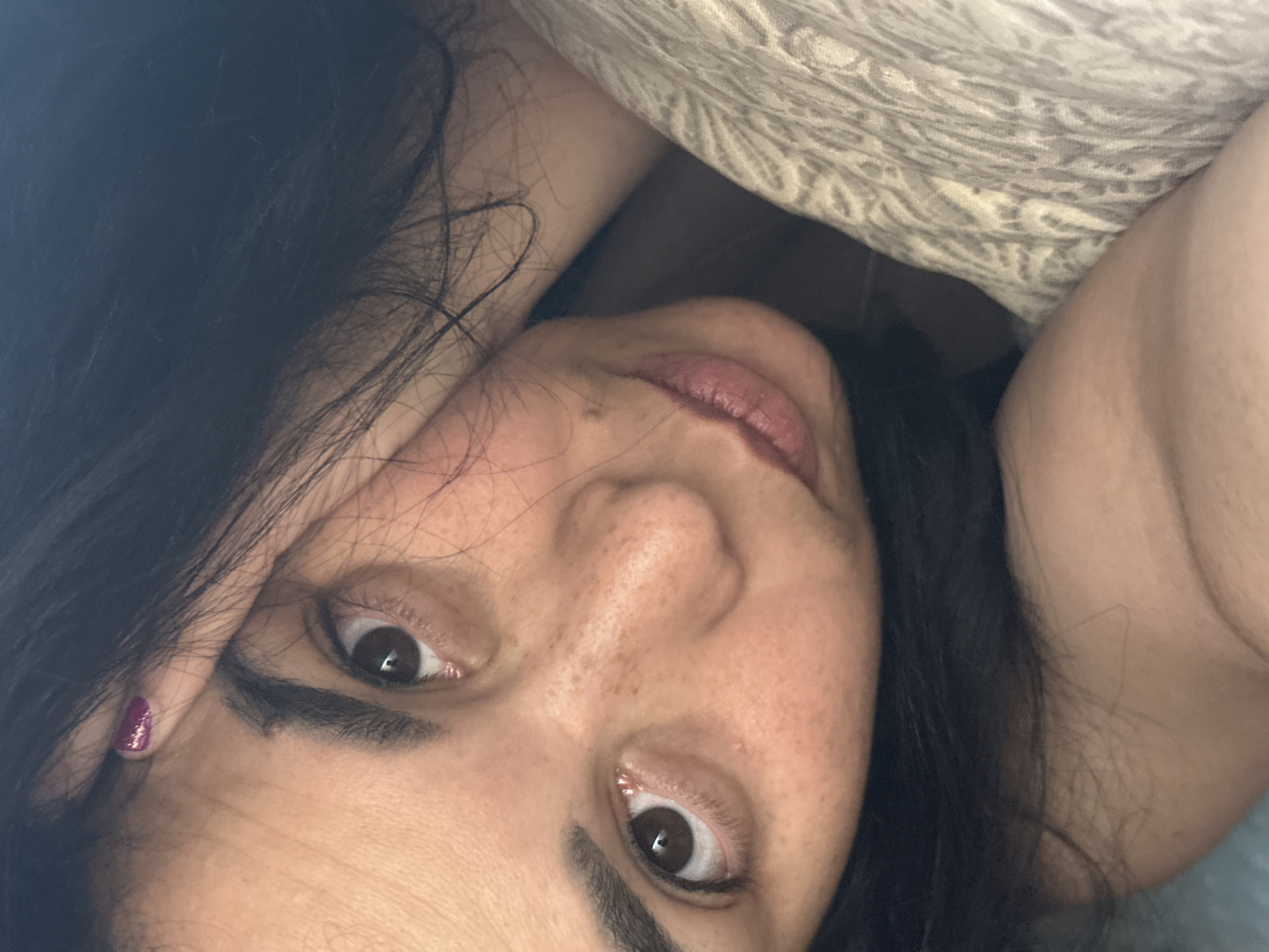 A close-up, overhead shot captures a young woman lying on a textured, light-colored blanket. Her dark hair is spread around her face, and her left arm is bent, with her hand resting on her cheek. The woman has brown eyes, dark eyebrows, and freckles across her nose and cheeks. Her lips are slightly parted, and a small mole is visible on her chin. She is wearing dark nail polish on her right hand, which is also resting near her face, partially obscuring her eye. The lighting is soft, and the focus is on her facial features.