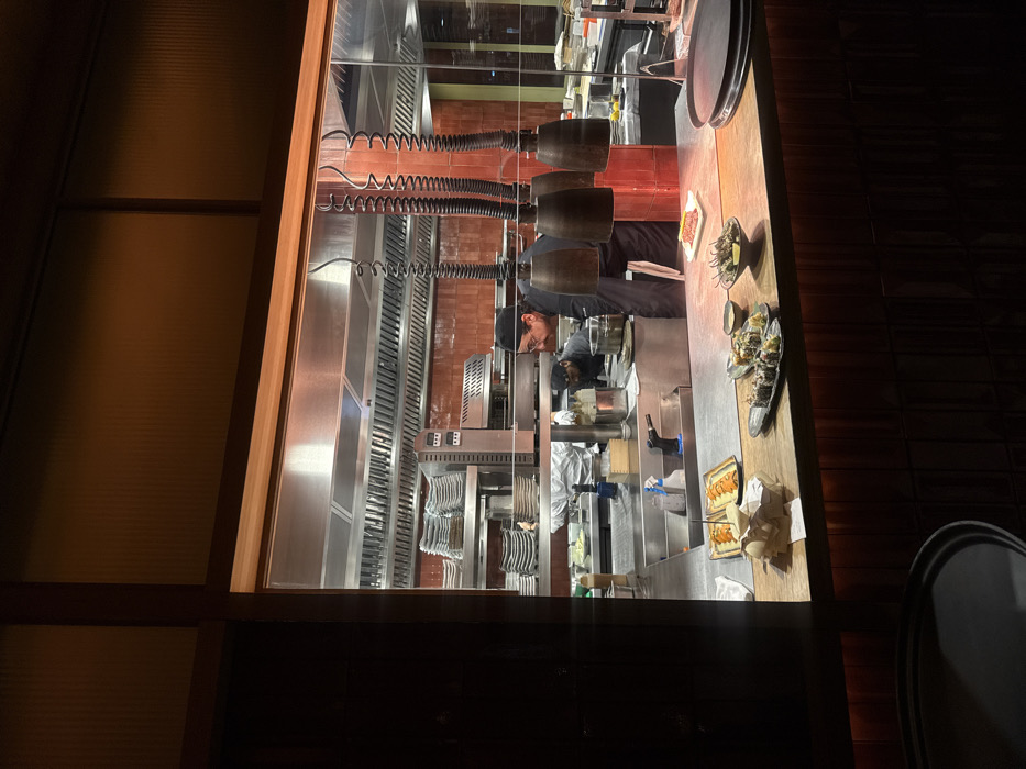 This image captures an active professional kitchen within a restaurant in Paris, France, viewed through a large, illuminated rectangular window from a darker dining area. Inside the kitchen, two male chefs are engaged in food preparation on stainless steel counters under extensive exhaust hoods. The chef in the foreground, wearing glasses and a black uniform, leans over a workstation, while another chef is partially visible behind him to the left. Stacks of clean white plates are arranged on a stainless steel rack to the left. The back wall of the kitchen is tiled in reddish-brown. On the wooden pass-through counter in the immediate kitchen foreground, several dishes are presented for service: two rectangular plates of sushi rolls with an orange topping, a longer black plate of assorted sushi, a black bowl containing green peppers, and a small white rectangular dish of red tartare. Four industrial heat lamps with coiled black cords hang directly over these prepared items. The foreground, from the viewer's perspective, is dark, showing the edge of a table or bar and a black circular tray or plate.