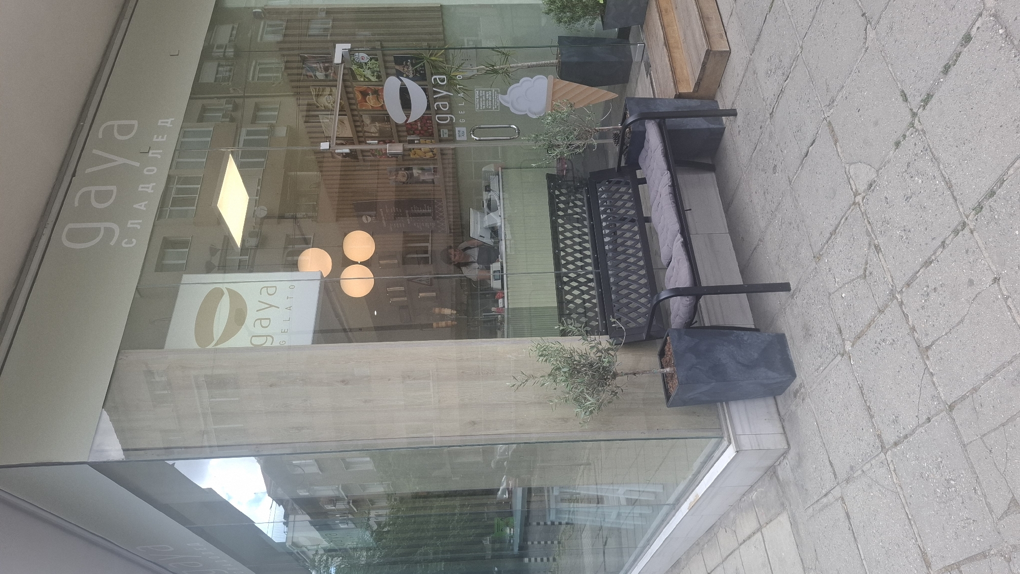 The image shows the exterior of a "Gaya Gelato" shop in Sofia, Bulgaria, as indicated by the Cyrillic text "СЛАДОЛЕД" (sladoled) meaning "ice cream" under the "Gaya" logo on the storefront. The shop has a large glass facade with the "Gaya Gelato" logo visible. Outside the shop, there is a black metal bench with grey cushions, two potted olive trees, and a wooden staircase leading to the entrance. A woman is visible behind the counter inside the shop, appearing to be working. The street outside is paved with concrete tiles.