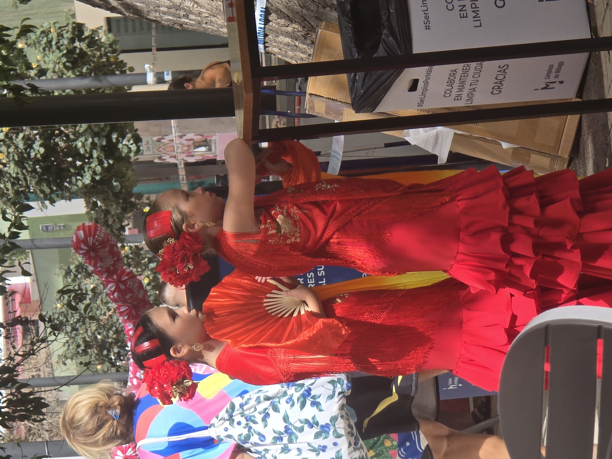 This image captures a vibrant street scene in Malaga, Spain, likely during a festival or cultural event. In the foreground, two young girls are dressed in traditional flamenco attire. The girl on the left wears a red dress with tiered ruffles and a fringed shawl, accessorized with a red flower in her hair and dangling earrings. She holds a bright orange fan. The girl on the right also sports a similar red flamenco dress with ruffles and a fringed overlay, adorned with a red flower hairpiece and earrings. Their dresses are richly detailed with fringe and embroidery.
Behind the girls, a woman with blonde hair is visible from the back, wearing a colorful, patterned top. In the background, trees with lush green foliage are abundant, suggesting an outdoor setting. A brown wooden table stands to the right, supported by dark metal legs, and a black trash bin with white text is placed beside it. The text on the bin appears to be a public service announcement, possibly related to cleanliness, with "#SerLimpiosPorMálaga" and "Limpieza de Málaga" visible, indicating the city of Malaga. The overall atmosphere is festive and bright, with a clear focus on the traditional Spanish cultural representation through the children's costumes.