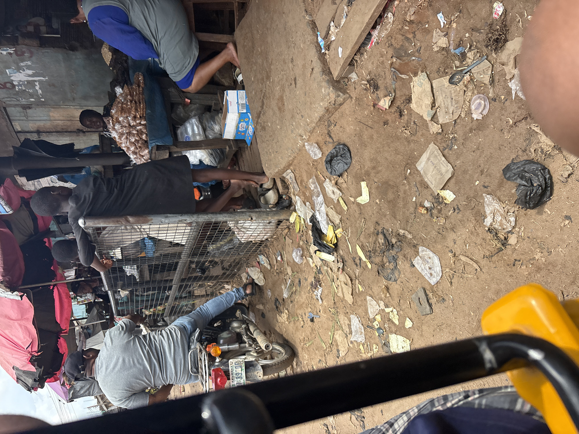 The image depicts a street scene in Fegge, Nigeria, likely a market area, characterized by an abundance of scattered debris, including plastic bags, paper scraps, and discarded food packaging, on the dusty ground. In the foreground, a person's legs and a portion of a yellow object, possibly part of a vehicle or a seat, are visible, suggesting the vantage point of a passenger.
Further into the scene, a man wearing a grey t-shirt and a black baseball cap sits on a motorcycle. He is facing away from the camera, towards a metal fence. Behind the fence, several individuals are engaged in activities related to commerce. One person, dressed in a black outfit, appears to be standing or leaning, while another, a woman with short hair, sits behind a table laden with bags of what seems to be food items, possibly nuts or grains. A third person, wearing a grey and blue garment, is seated on a stool to the right, facing the vendor.
The background shows makeshift stalls with umbrellas, indicating a market environment. The overall impression is one of a busy, possibly informal, marketplace with a notable lack of cleanliness due to the widespread litter.