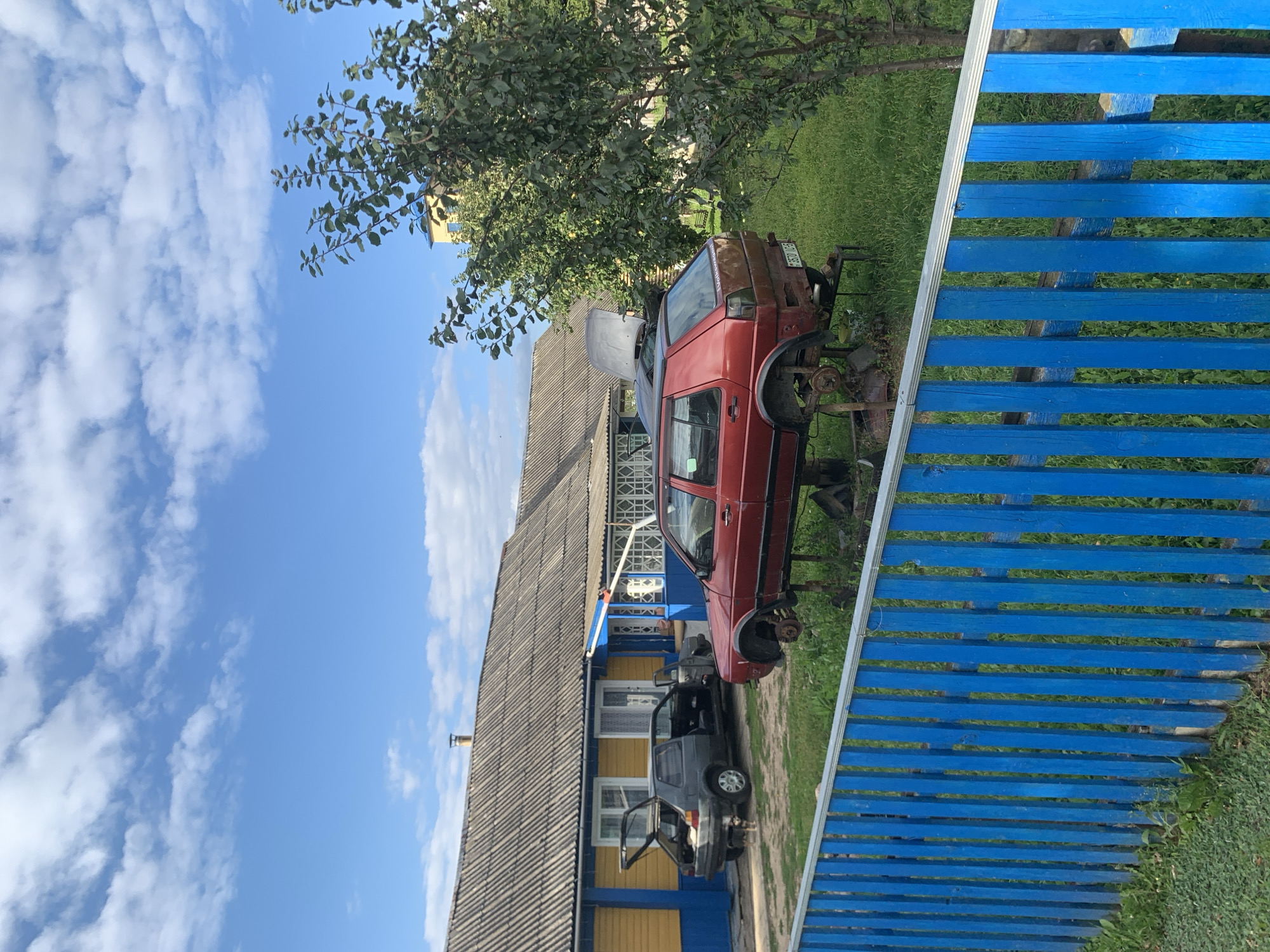 In the city of Kapyĺ, Belarus, a rural scene unfolds with two vehicles in a state of disrepair. In the foreground, a vibrant blue picket fence borders a grassy yard. Behind it, a red hatchback car is elevated on metal stands, its wheels removed, suggesting it's undergoing maintenance or is being dismantled. To the left, a dark gray hatchback is parked with its doors and trunk open, also appearing abandoned or under repair. A small dog stands near the gray car. A low-slung building with a grey, corrugated roof and yellow and blue siding dominates the background, with a visible chimney. A large, leafy tree stands to the right, partially obscuring a taller, light-yellow building in the distance. The sky is a clear blue with scattered white clouds.