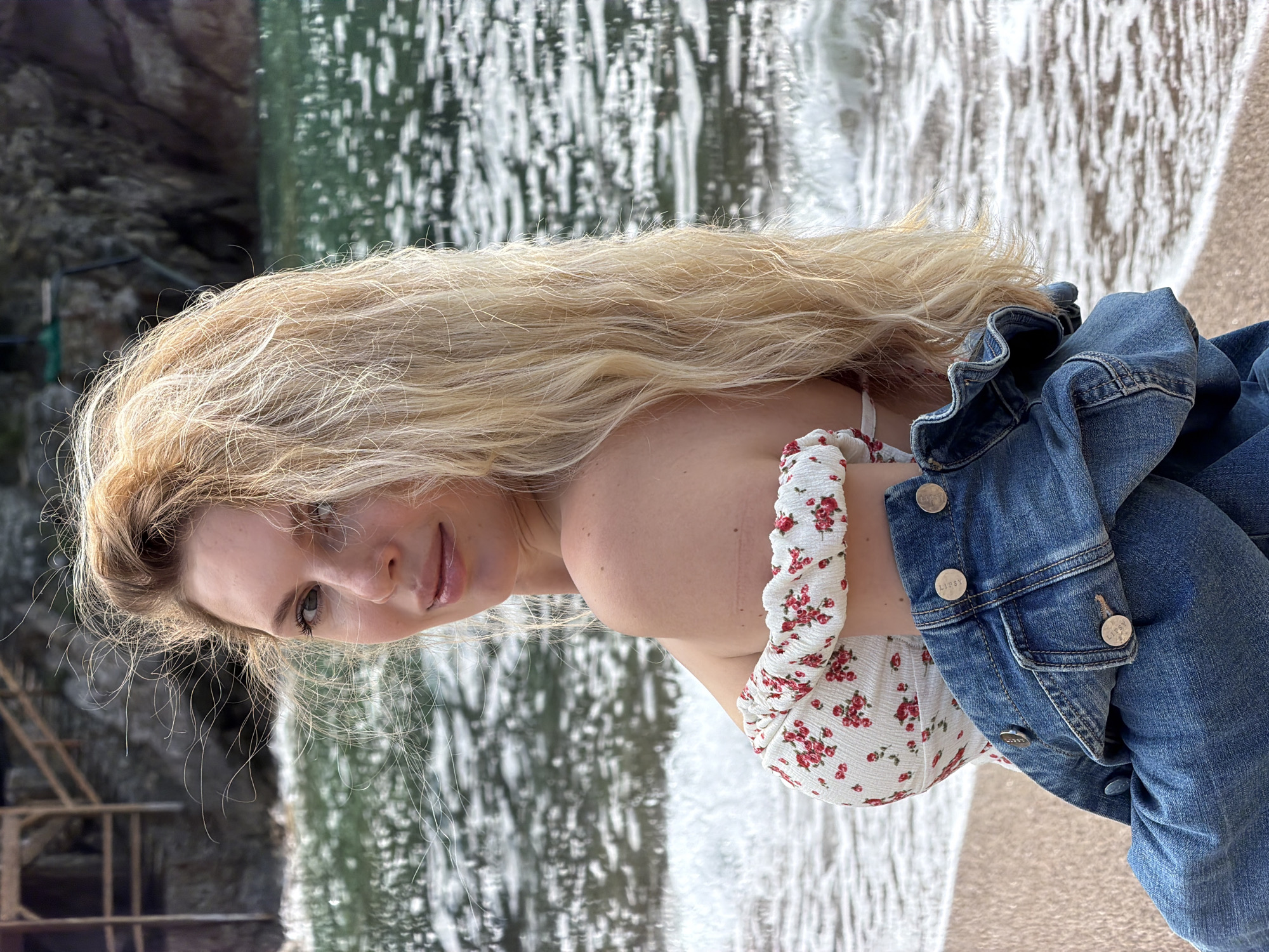 A woman with long blonde hair is at the beach near Petrovac centar, Montenegro, wearing a white floral top and a blue denim jacket. The location includes a beach and the sea with the beach occupying the bottom right of the frame and the sea behind the woman. A rocky outcropping on the left suggests the coastal nature of Petrovac centar.