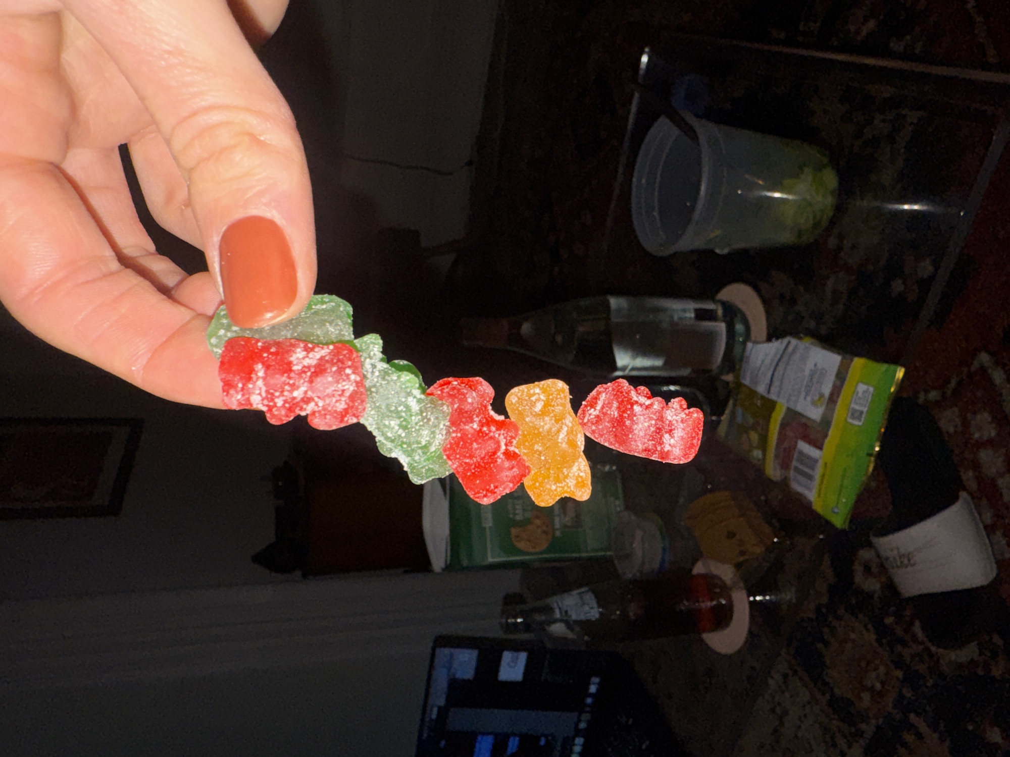 A person's hand holds a string of gummy bears (green, red, orange, and red) over a cluttered glass coffee table. On the table, there is a laptop, a cup, a wine bottle, a snack bag, and a cookie box. A pair of sandals and a framed painting can be seen in the background. The location cannot be determined from the image.