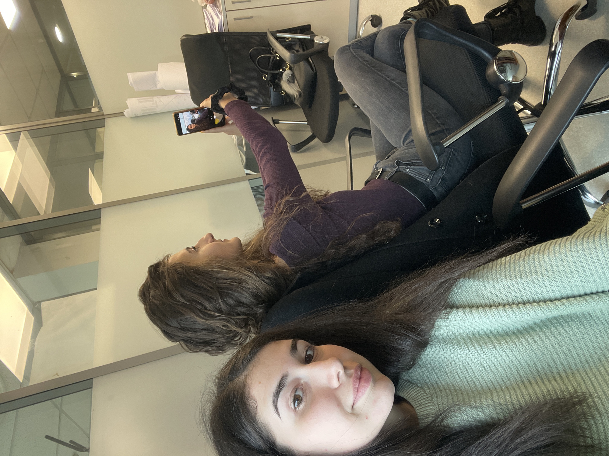 Two young women are in a modern office space. One woman, wearing a light green sweater, is in the foreground. Behind her, another woman in a purple top lies back and holds up a phone, seemingly taking a picture or video. The location is likely Sofia, Bulgaria. Office furniture includes office chairs, a cabinet, and documents. The environment suggests a workplace or a break area in an office.