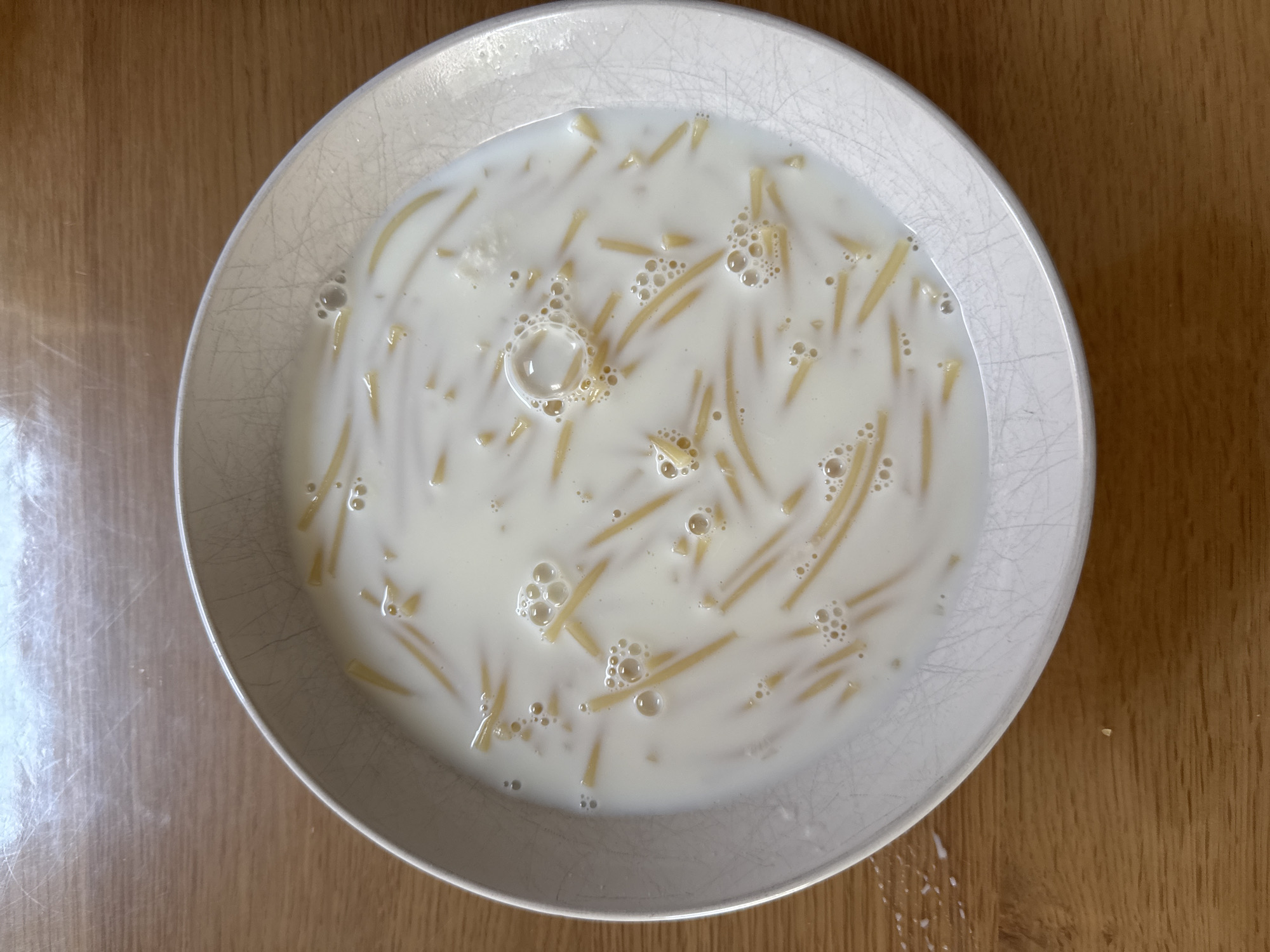 A white, rimmed bowl filled with milk and vermicelli noodles sits on a wooden surface. The milk is opaque white, and the thin, golden noodles are submerged in it. Bubbles are visible on the surface of the milk, indicating it may be warm or freshly poured. A few splashes of milk are visible on the wooden table to the left of the bowl. The wooden surface has a visible grain and appears to be a table. The lighting suggests an indoor setting.