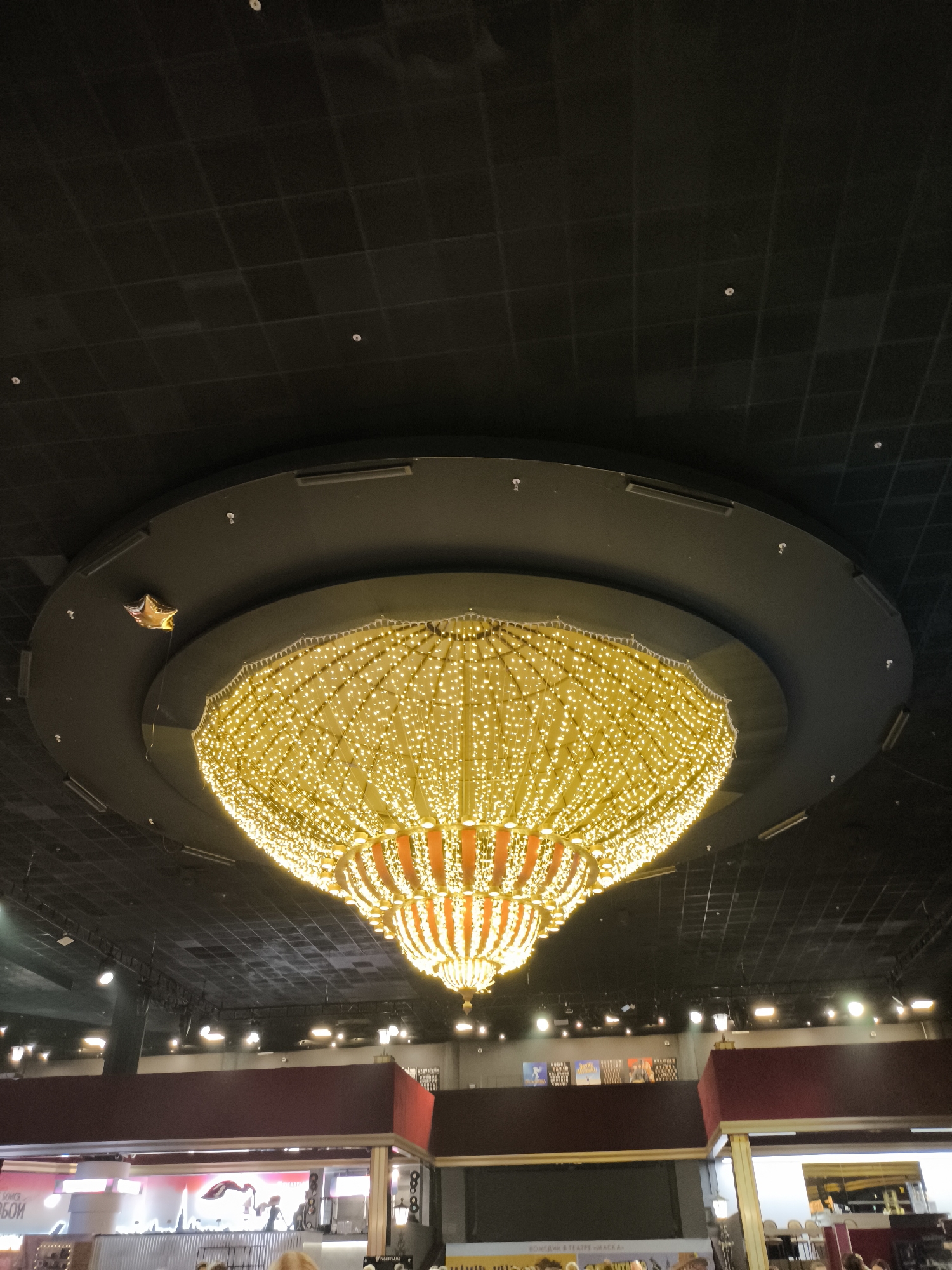 An indoor shot of a large, ornate chandelier illuminated with many small, bright yellow lights, hanging from a dark, coffered ceiling. The chandelier is dome-shaped and has multiple tiers, with some orange elements visible in its structure. A single golden star-shaped balloon is attached by a string to the dark ceiling above and to the left of the chandelier. Below the chandelier, partially visible, are the tops of some structures with maroon colored panels and some lighting. The scene appears to be within a public building, possibly a theater or entertainment venue, based on the scale and decor.