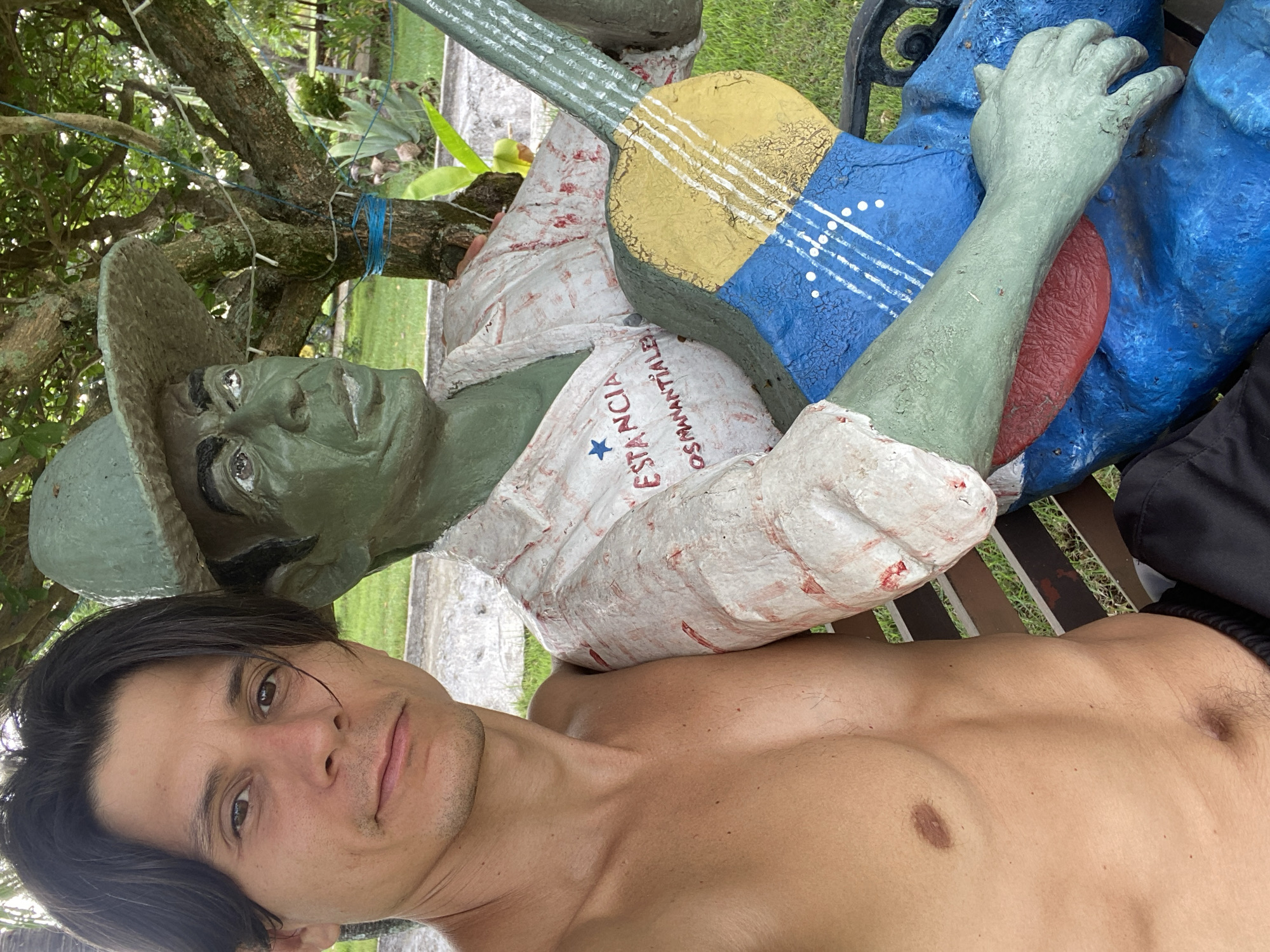 An outdoor portrait captures a man taking a selfie with a distinct statue. The image is oriented sideways, with the man's head on the left. The man, with dark hair and a neutral expression, is shirtless, revealing his torso. He is positioned alongside a prominent statue of a man.
The statue features a green-skinned figure wearing a light-colored, long-sleeved shirt and a wide-brimmed hat. The shirt has red lettering, including "ESTANCIA" and "OSAMANTHA LEI," along with a blue star. The statue is holding a guitar that is painted with the colors of the Venezuelan flag: yellow, blue, and red, with white lines and dots representing strings and stars on the blue section. One of the statue's green hands is visible resting on a blue object, possibly part of another statue or a bench.
The setting appears to be a lush, green outdoor area, likely a garden or yard, with trees and grass visible in the background, along with a paved surface. The lighting suggests it is daytime. The presence of the Venezuelan flag on the guitar, combined with the "Vallecito, Venezuela" location context, strongly indicates the image was taken in Venezuela, possibly at an estate or public art display.