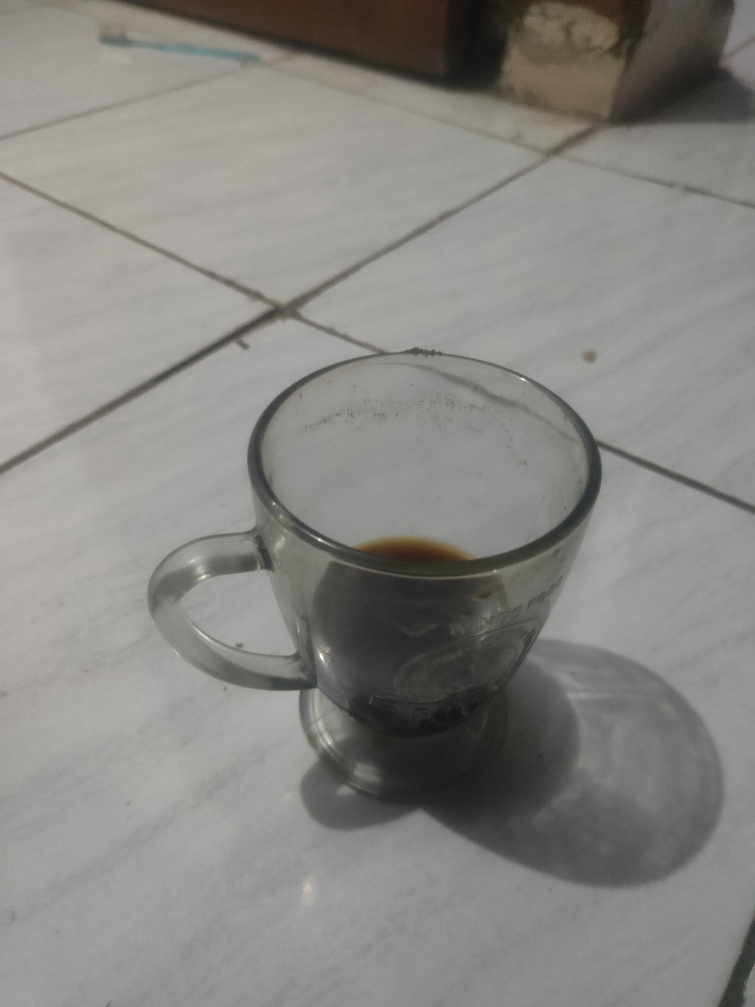 A close-up, slightly blurry, overhead shot shows a glass cup with a handle, filled with a dark liquid, possibly coffee, sitting on a tiled surface. The tiles are light-colored with dark grout lines, forming a grid pattern. In the background, out of focus, is a wooden surface and a light-colored object. The lighting suggests an indoor setting, possibly a home in Antirogo, Indonesia, with a shadow cast by the cup on the tiles.