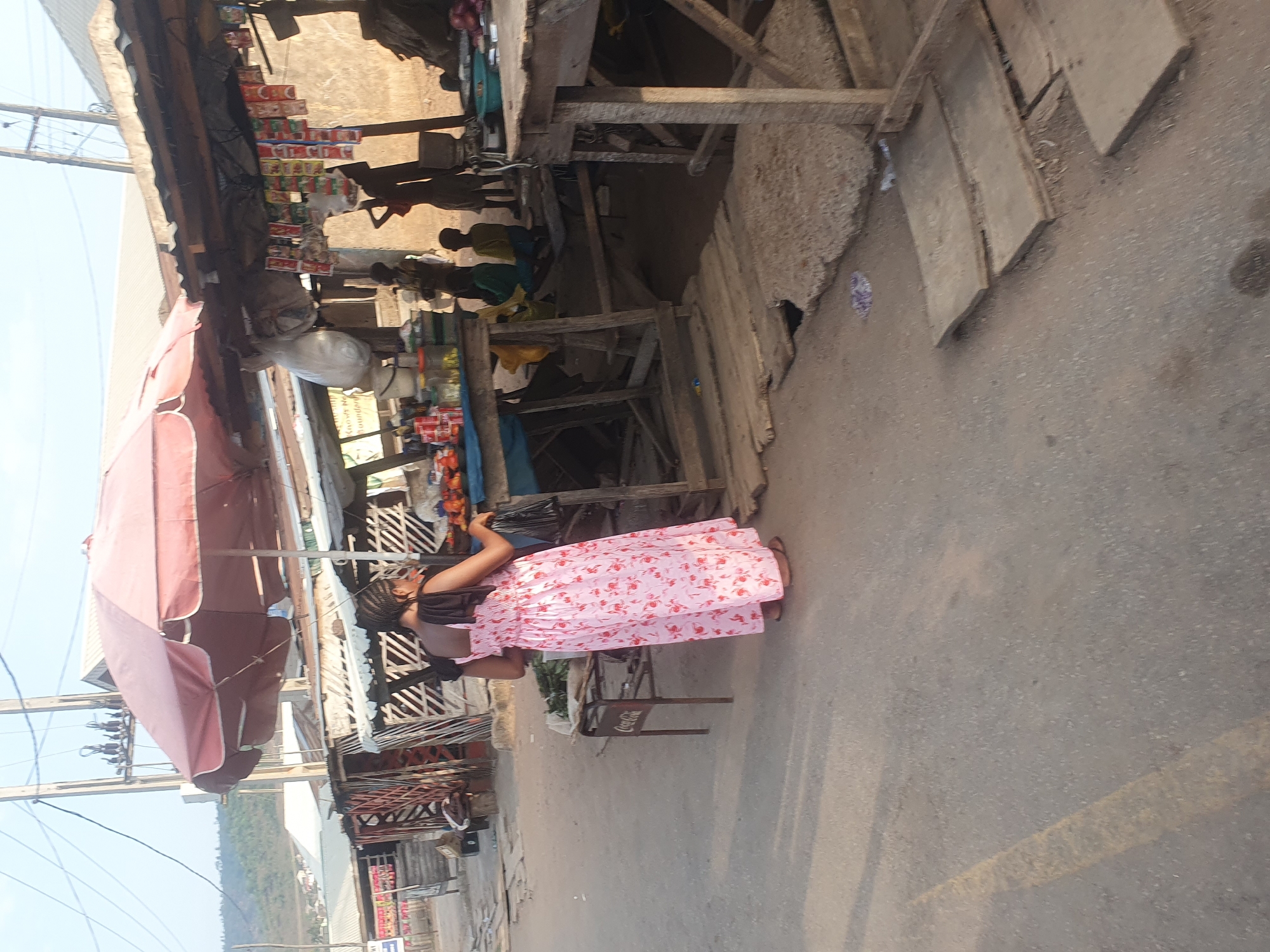 The image shows a street scene in Lokoja, Nigeria. A woman in a pink patterned dress stands in the street near a market stall. The market stall is covered by a large pink umbrella. Other market stalls are seen in the background. People are present near the stalls. Overhead power lines are visible. The street is paved and appears to be in a residential or commercial area.