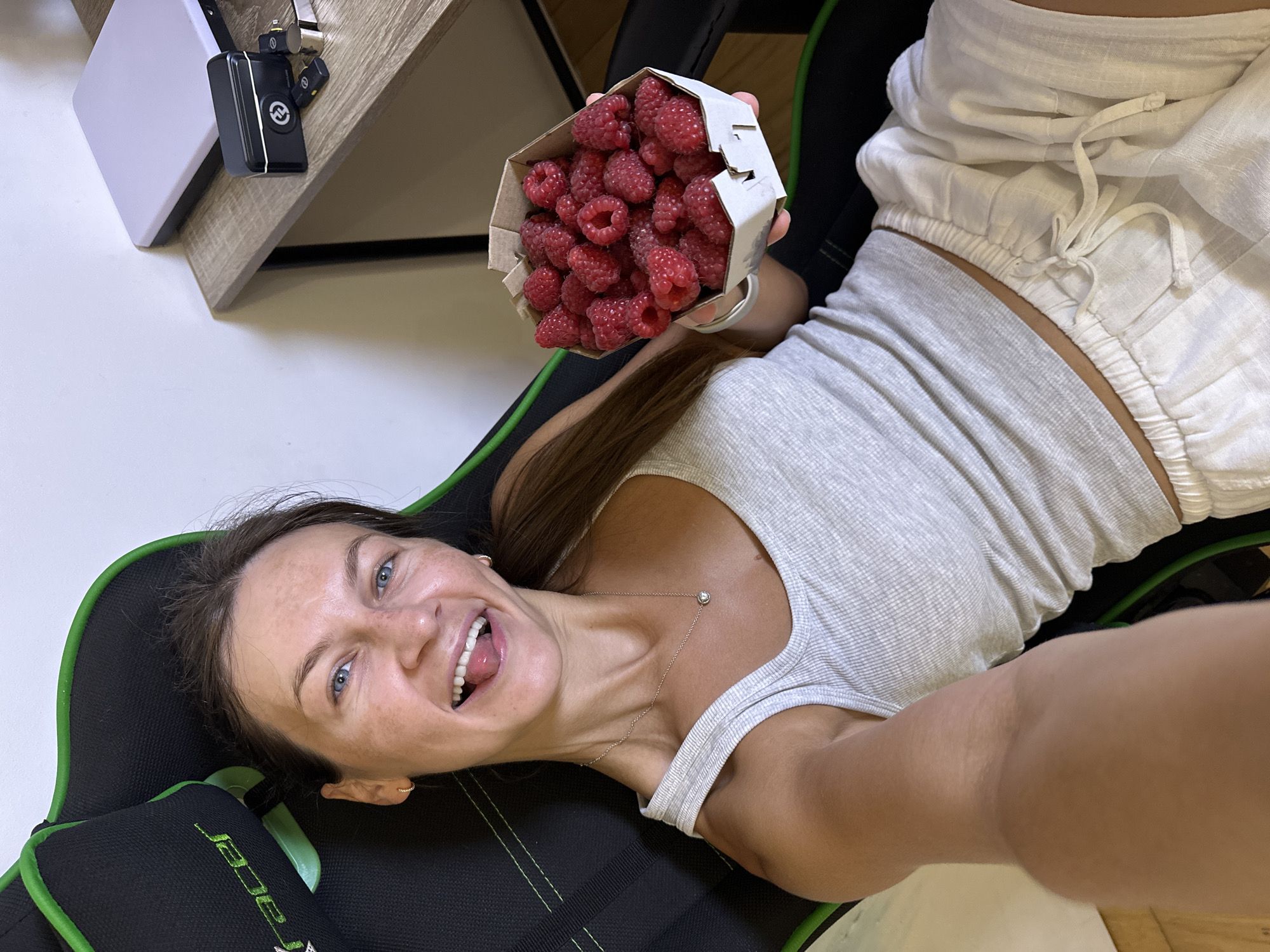 A woman is seated in a black and green gaming chair, holding a cardboard container filled with raspberries. She is looking at the camera and sticking out her tongue playfully. The woman is wearing a light grey tank top and white shorts. To her right, a wooden desk is visible with a laptop and some small electronic devices. The background appears to be an indoor setting with a white wall. The specific location in Boreti, Montenegro is not discernible from the image.