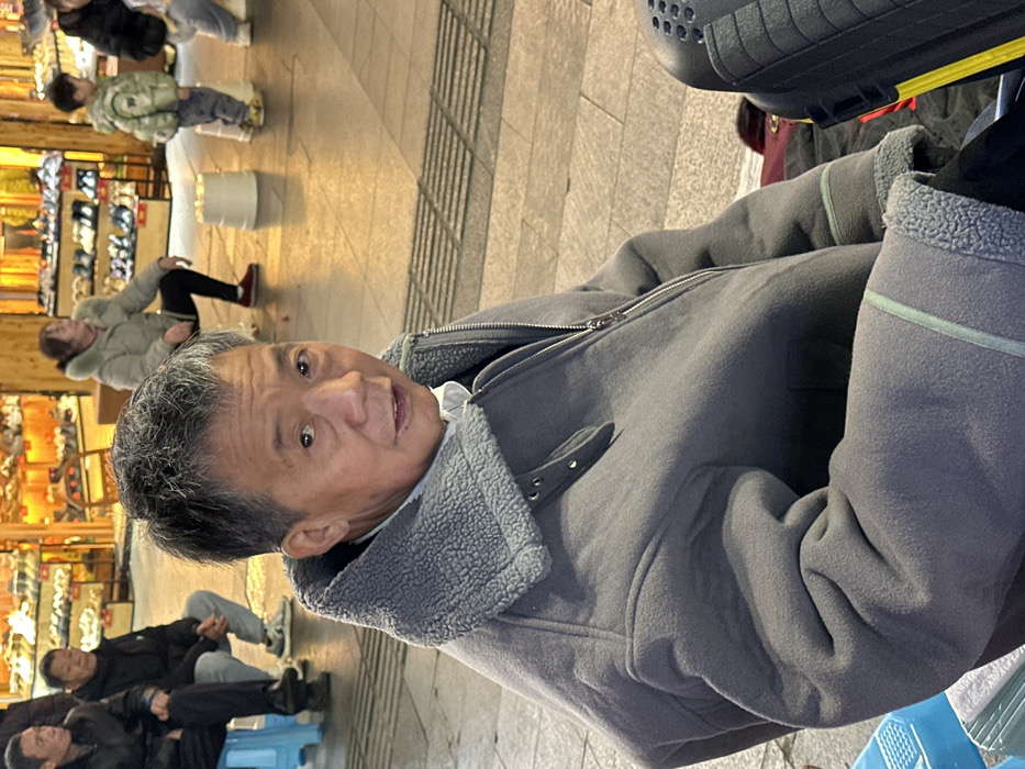The image depicts a bustling commercial pedestrian area in Jiangjin, China. In the foreground, an adult Asian male is seated, facing the viewer directly with a slightly open mouth. He wears a grey jacket with a prominent grey faux sheepskin collar and lining visible at the cuffs, over a white shirt. To his left, a light blue plastic stool is partially visible, and to his right, a black object with a perforated top surface.
In the mid-ground to the left, two other adult Asian males are seated on light blue plastic stools. One wears a dark jacket and dark pants, while the other is dressed in a dark jacket, light jeans, and sneakers. To the right, a woman in a light puffer jacket, dark pants, and red shoes squats with her back partially to the viewer. A small child in a light green puffer jacket stands behind her. Two small white cylindrical objects rest on the paved ground nearby.
The background features brightly illuminated retail displays showcasing numerous pairs of shoes on shelves. The ground is paved with light-colored tiles, including a section of grey tactile paving with parallel ridges.