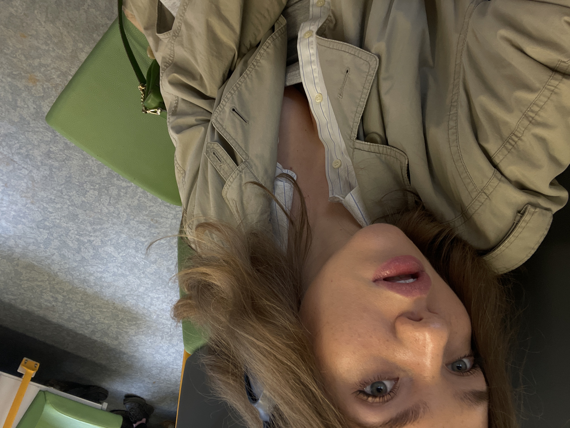 A person is viewed from a slightly high angle, facing the camera. They have light brown hair, fair skin, and light-colored eyes. They are wearing a beige-colored jacket over what appears to be a light-colored collared shirt. A green purse is positioned to the left of the person. The background includes a gray floor. The location of the city of Moscow, Russia is not discernible from the image.
