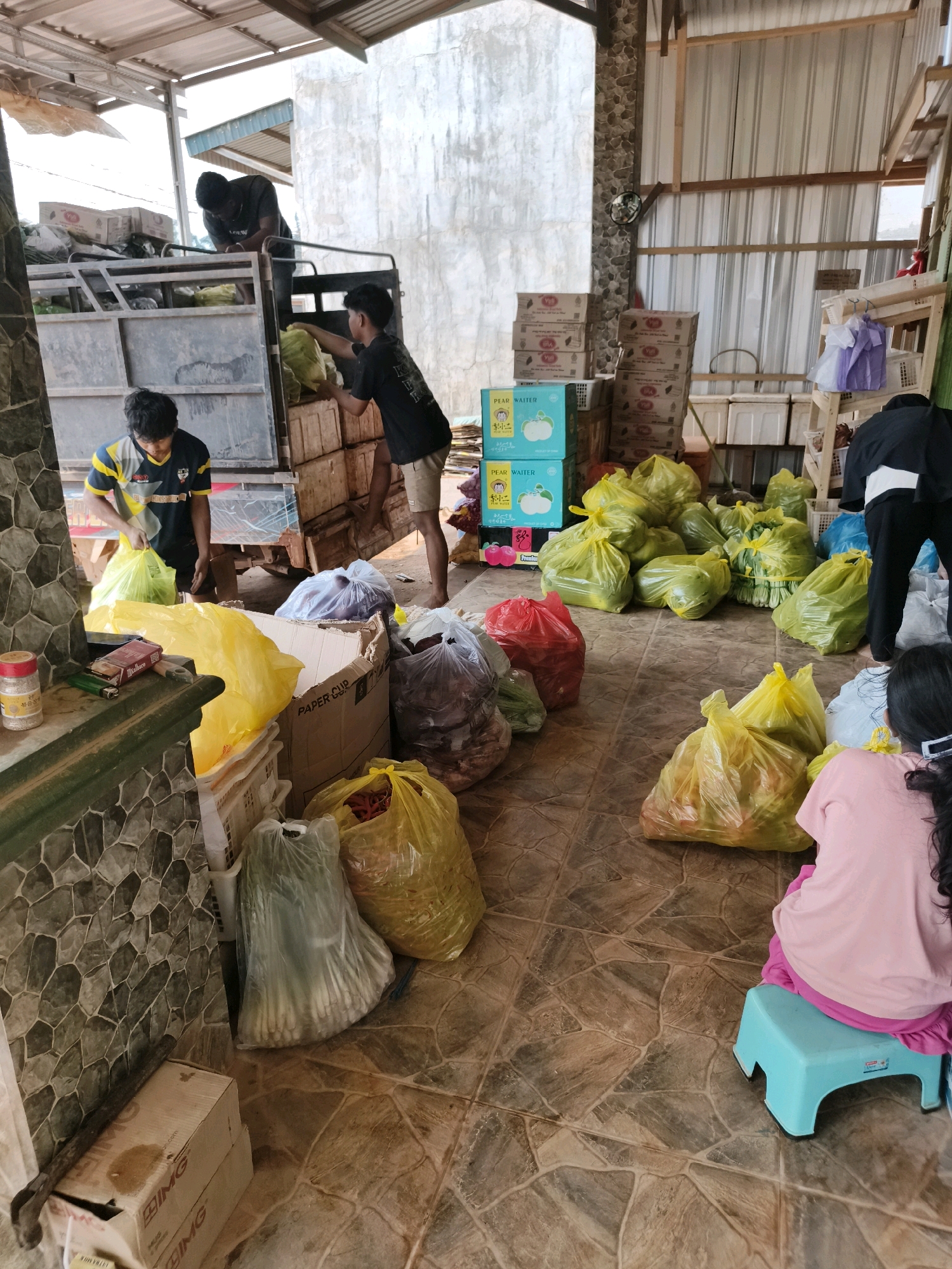 The image depicts a scene within a covered outdoor market or storage area, possibly in Ketapang, Indonesia. In the background, a truck is being loaded with what appear to be produce items. Three individuals are present; two are actively engaged in the loading process, while a third, partially visible, is also involved.
The foreground is cluttered with numerous large, transparent yellow plastic bags, presumably filled with fruits or vegetables. Other bags, some red and some opaque white, are also visible, containing various items. Stacked cardboard boxes and white plastic crates are scattered throughout the space. A child is seated on a small blue plastic stool, facing away from the camera, wearing a pink shirt and dark pants.
The environment is characterized by a rustic, open-air structure with a corrugated metal roof and partially constructed walls made of stone and concrete. The flooring appears to be tiled with a stone-like pattern. The overall impression is one of a busy, operational area for the distribution or storage of goods.