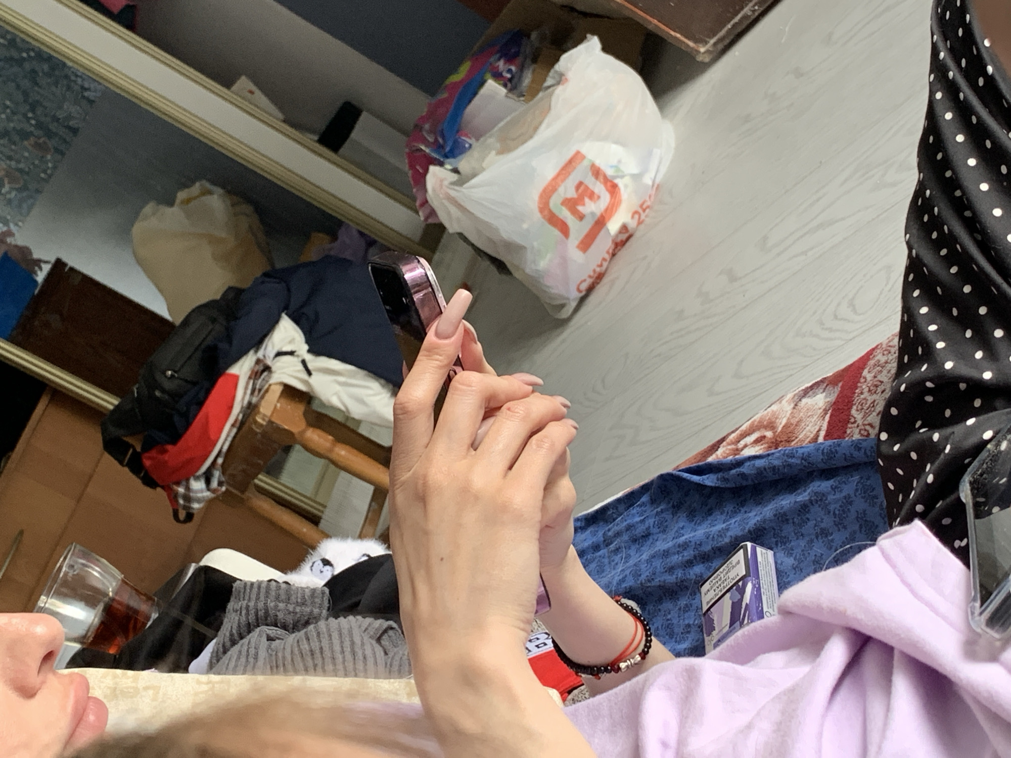 Scene: A person is lying down, holding a mobile phone with very long, manicured fingernails.
People: One person is visible, lying down and holding a phone. Only part of their face is visible.
Objects: Mobile phone, long manicured fingernails, bracelet, glass of liquid, clothes, white grocery bag, cigarettes, bedsheets.
Actions: The person is holding a mobile phone.
Environment: The scene appears to be indoors, possibly in a bedroom or living space. Clothes are piled nearby, along with a glass of liquid.
Location: Stavropol, Russia.