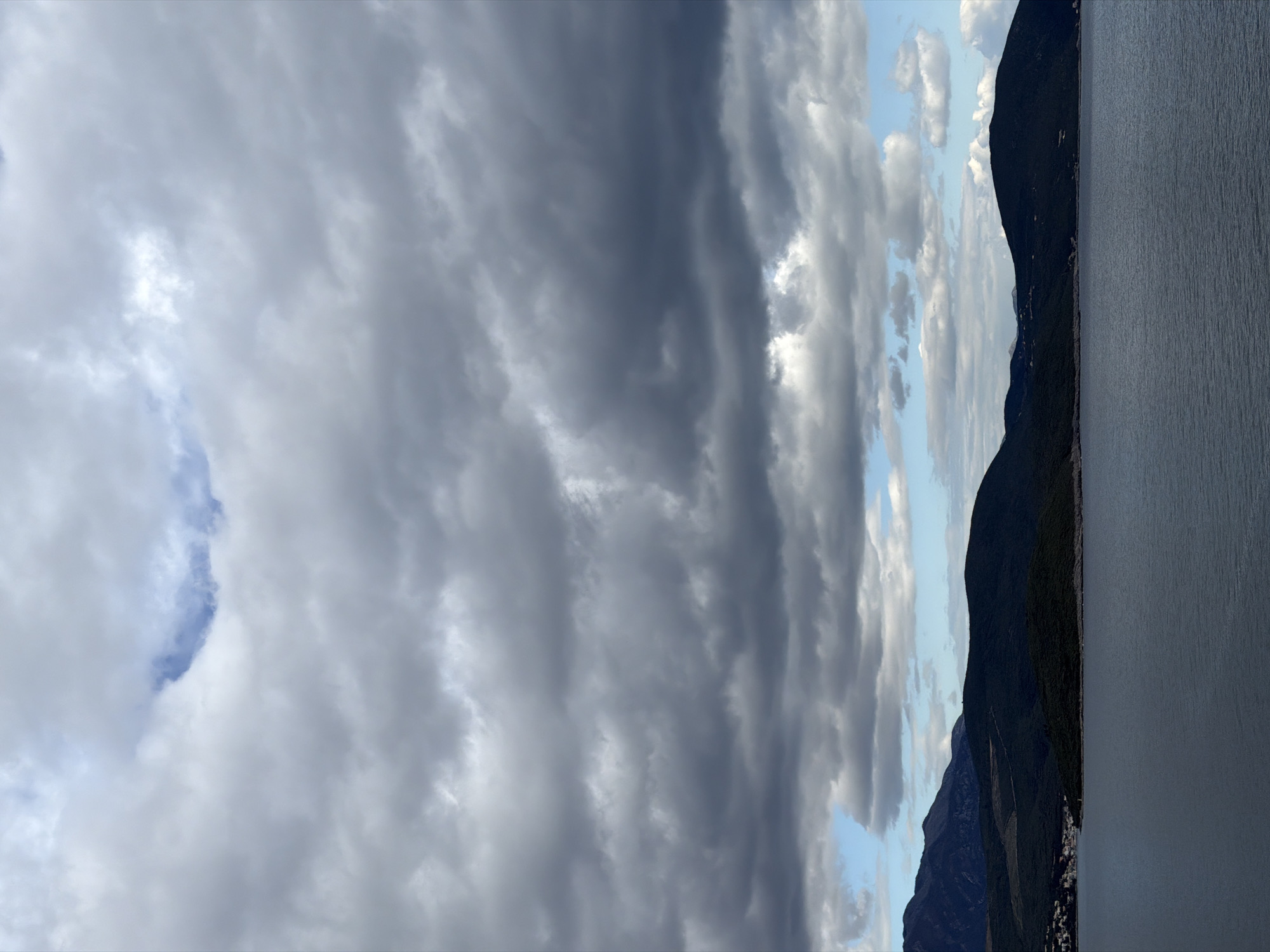 The image shows a landscape in Radovići, Montenegro. Dark mountains rise along the right side of the frame, bordering a body of water. The sky is filled with varying cloud formations, ranging from dense, dark gray clouds to lighter, patchy clouds with glimpses of blue sky.
