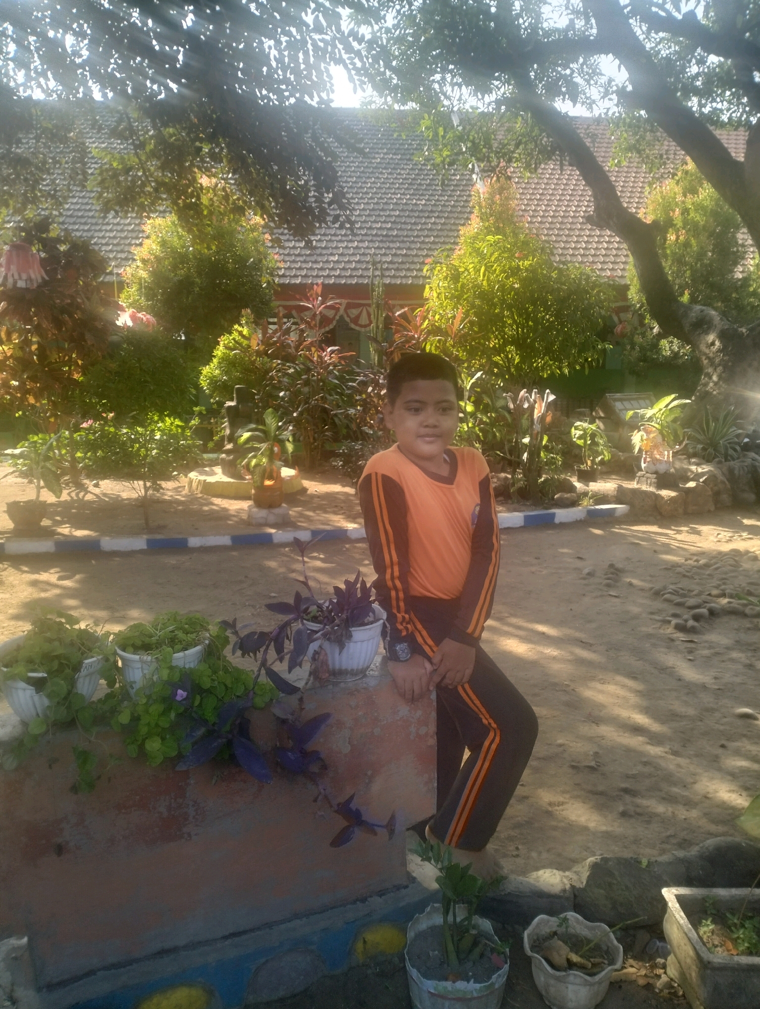 A young boy in an orange and black long-sleeved shirt and dark pants with orange stripes stands in an outdoor area with a tiled roof building and lush greenery in the background. He is leaning on a low wall. Several potted plants are visible in the foreground, and the ground is covered in dirt and small stones. The scene appears to be in Blabakan, Indonesia, given the architectural style and vegetation. The lighting suggests it is daytime.