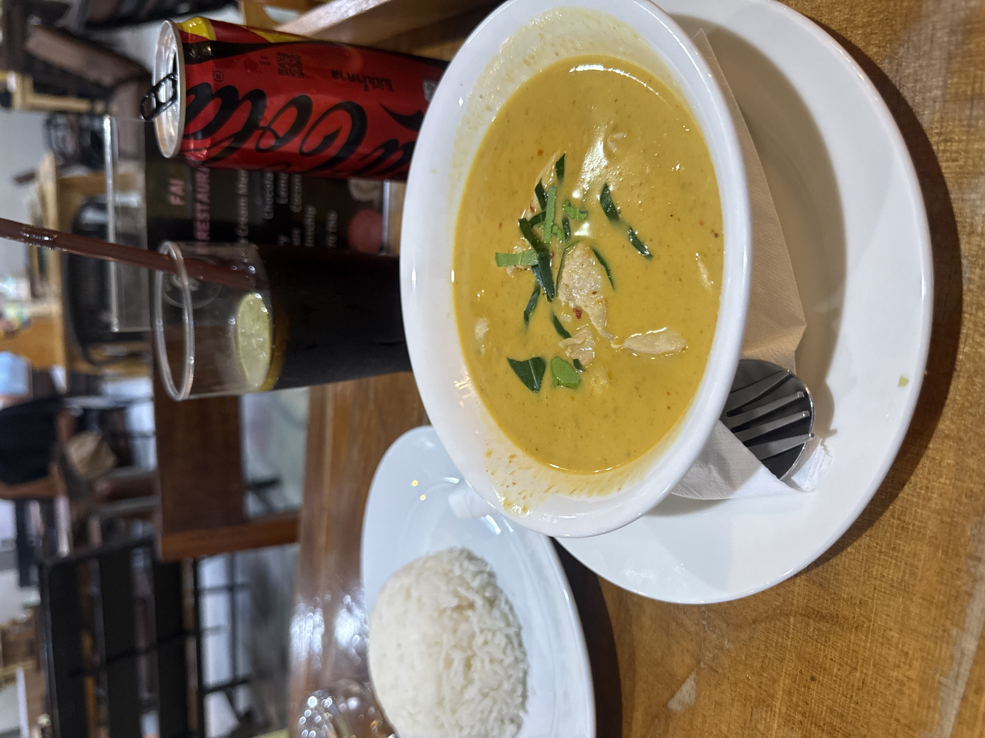 A close-up, high-angle shot shows a meal on a wooden table. The meal includes a bowl of yellow curry with green garnishes and a side of white rice. To the left of the curry, a glass with a dark beverage and a straw is visible, next to a red Coca-Cola can. The scene appears to be an indoor setting, possibly a restaurant. The lighting suggests it could be daytime or evening. No people are visible in the frame. The background shows a blurred view of what appears to be a restaurant interior. Visible text on the Coca-Cola can reads "Coca-Cola." The overall impression is of a casual dining experience.