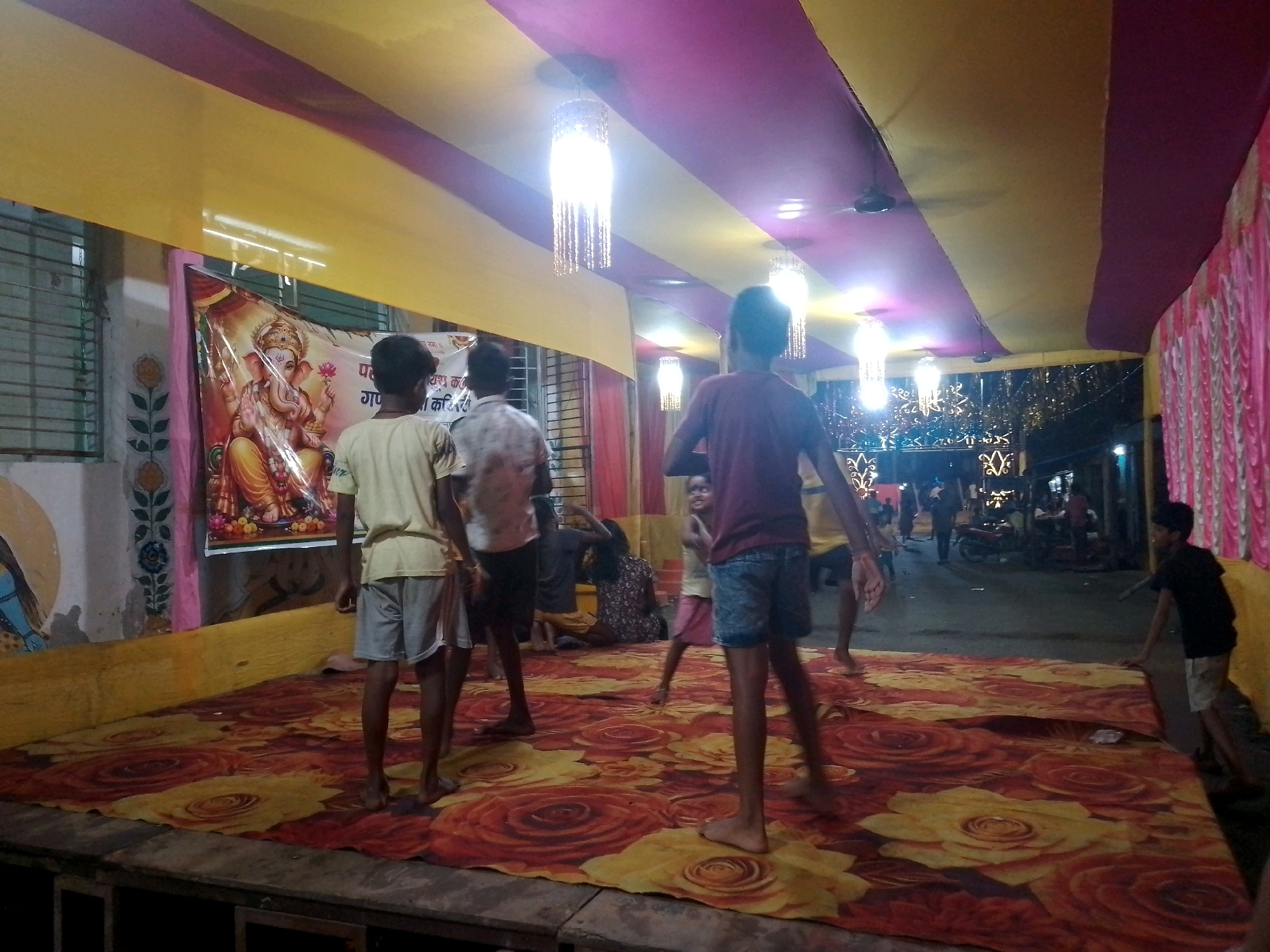 The image captures an outdoor event, possibly a festival or celebration, taking place under a temporary canopy structure in Siliguri, India. The canopy is draped with alternating yellow and purple fabric, and illuminated by hanging decorative lights and ceiling fans. In the foreground, a group of children and young adults are present. A large floral-patterned rug covers the ground in the immediate area, suggesting a stage or performance space.
To the left, a poster or banner featuring an image of the Hindu deity Ganesha is prominently displayed. Several individuals are standing on the rug, some facing the viewer and others engaged in activity. One young person in the foreground is wearing a yellow t-shirt and grey shorts. Further back, other children are visible, one in a maroon t-shirt and blue shorts, and another girl wearing a pink skirt and a light-colored top.
The background reveals a street scene with more people, motorcycles, and buildings, indicating an urban setting. The overall atmosphere appears lively and festive. The presence of the Ganesha image suggests a religious or cultural event, likely Hindu. The temporary structure and decorations point towards a community gathering.