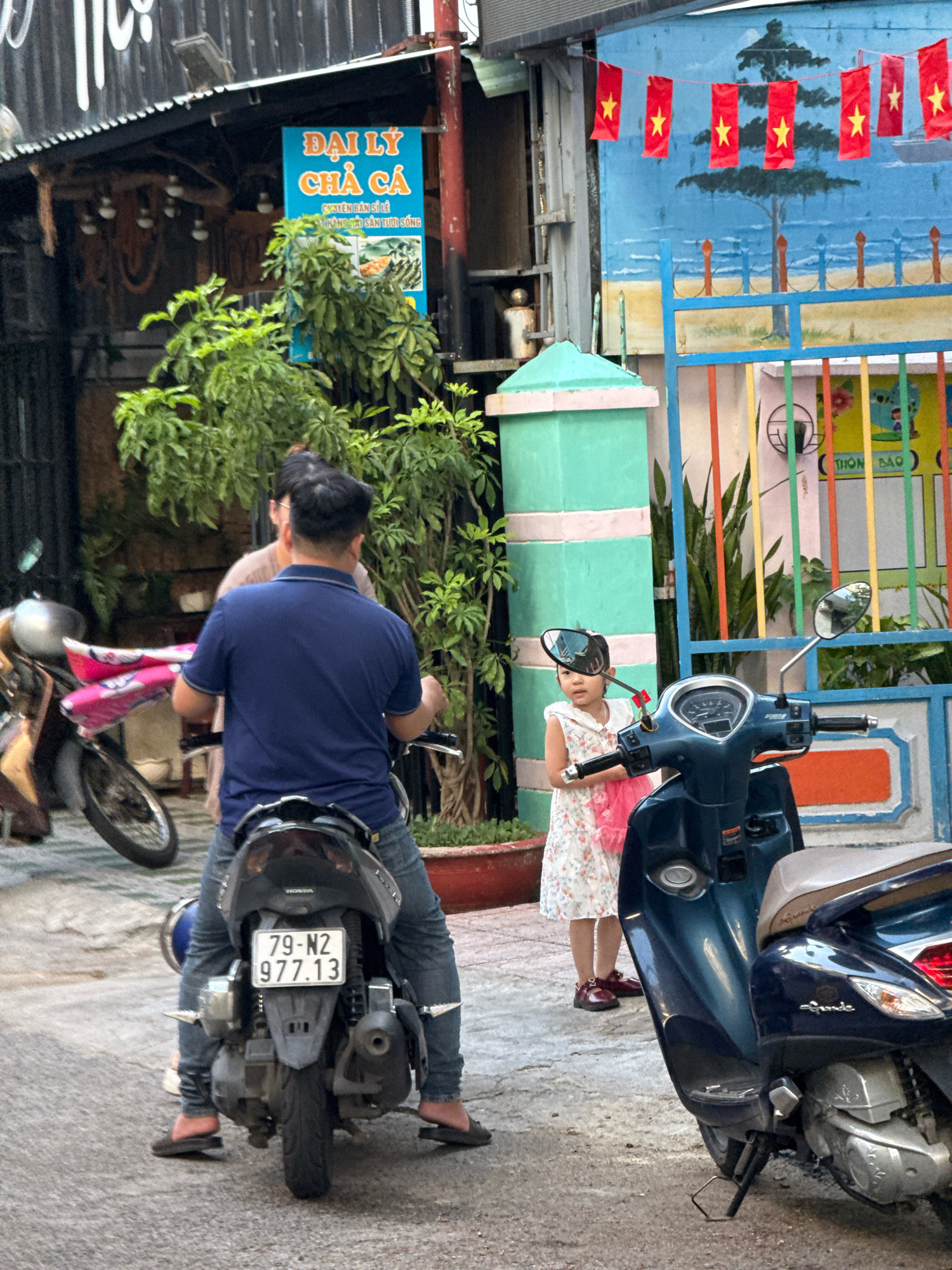 The image depicts a street scene in Nha Trang, Vietnam. In the foreground, a person is seated on a dark blue scooter, facing away from the camera. The scooter has a license plate with the number "79-N2 977.13". To the left of this scooter, another scooter is partially visible.
To the right of the first scooter, a young girl in a white floral dress and a pink sash stands near a light blue and pink segmented pillar. She is looking towards the viewer. Behind the girl, a dark blue scooter with a visible dashboard is parked.
In the background, there are buildings, including a shop with a blue sign that reads "ĐẠI LÝ CHẢ CÁ" (Fish Cake Agency) and some plants. Red and yellow Vietnamese flags are strung across a mural painted with a beach scene and trees. The environment appears to be an urban street setting with pavement and a building facade.
