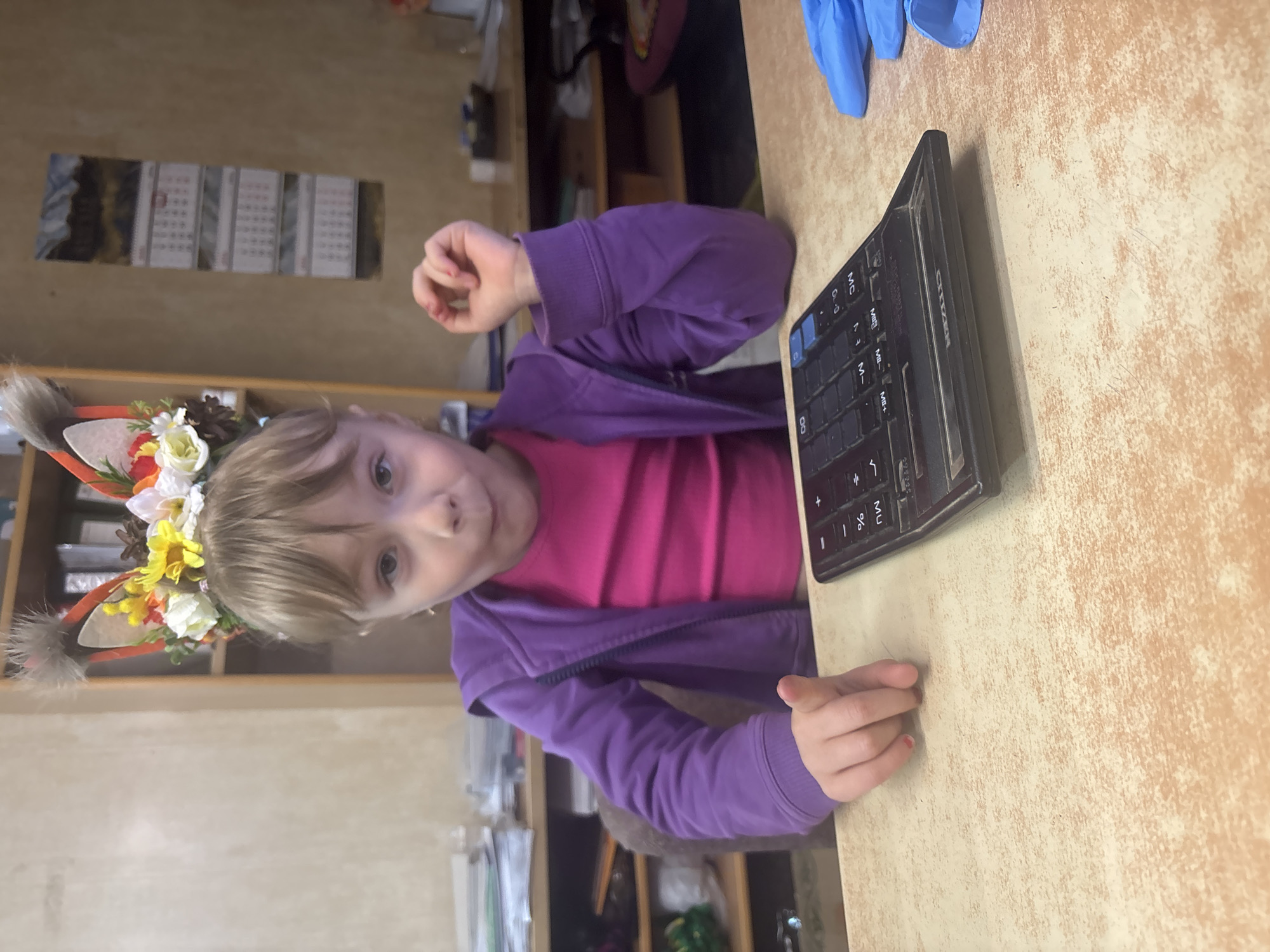 A young child with blonde hair and light-colored eyes, wearing a purple zip-up jacket over a pink t-shirt, is seated at a light brown desk or table in an indoor setting. She wears a decorative headband with faux animal ears, adorned with small white, yellow, and orange flowers, and pinecones. The child looks directly at the camera with a neutral expression.
On the desk to her right, a black, somewhat dusty "CITIZEN" brand calculator is visible, showing various function keys. Behind the calculator, a pair of blue disposable gloves is resting on the table. The child's right hand is partially closed, and her left hand rests on the table, with a hint of red on one fingernail.
The background shows a wall with a calendar featuring multiple months. Further back, shelves and other household or office items are blurred, suggesting a cluttered but functional space. The lighting appears to be ambient, likely from an overhead source or a window. The scene has a casual, domestic, or informal office feel. While the location context is Chaikovsky, Russia, there are no specific visual cues within the image to confirm this directly.