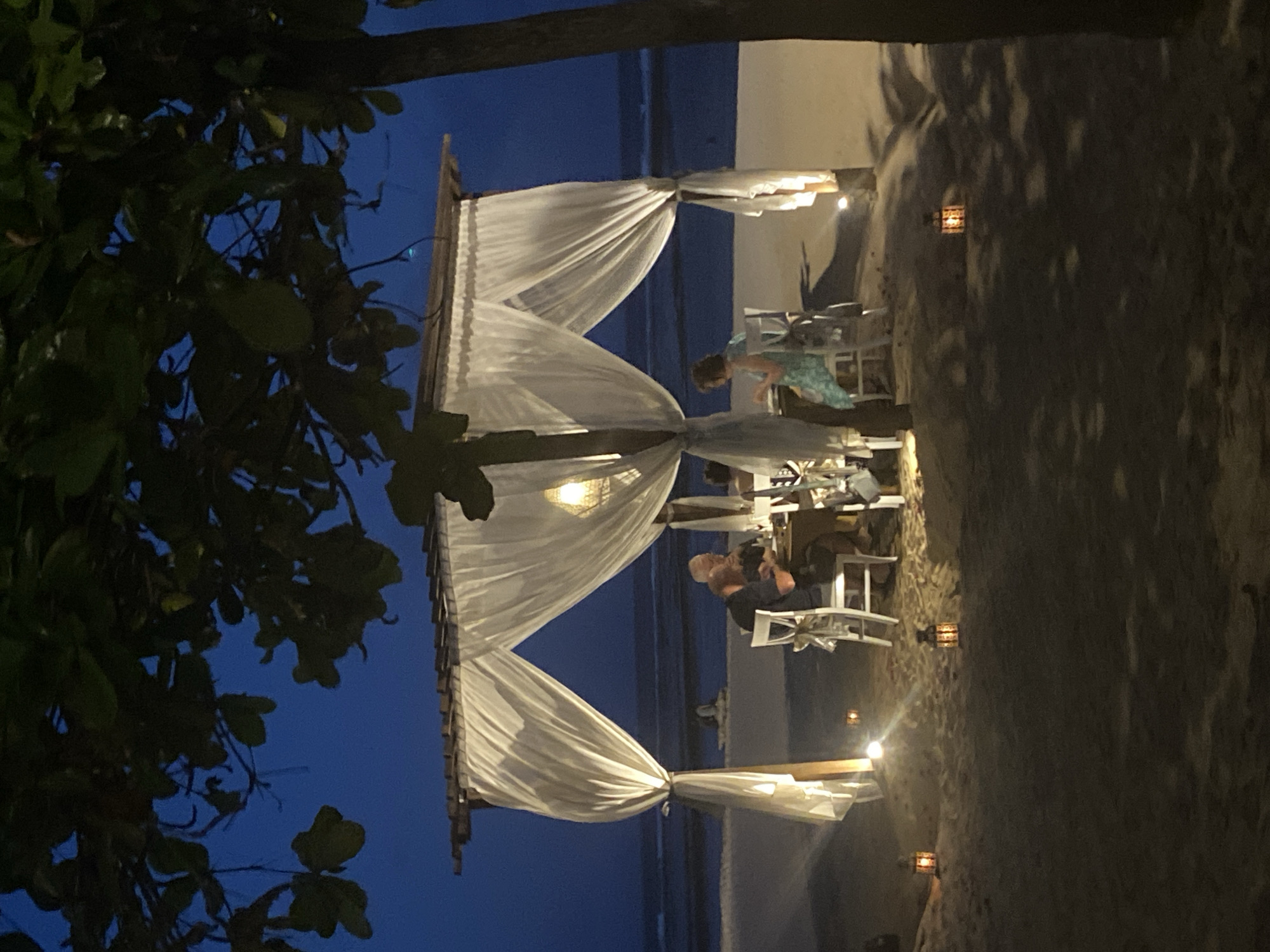 The image depicts an outdoor dining area at dusk in Kuta Selatan, Indonesia, likely on a beach. A canopy draped with white fabric covers a set of tables and chairs. Two people are seated at one of the tables, while another person stands nearby. Lanterns provide ambient lighting. A tree is visible in the foreground, obscuring part of the view. The beach and ocean are visible in the background, under a darkening sky.