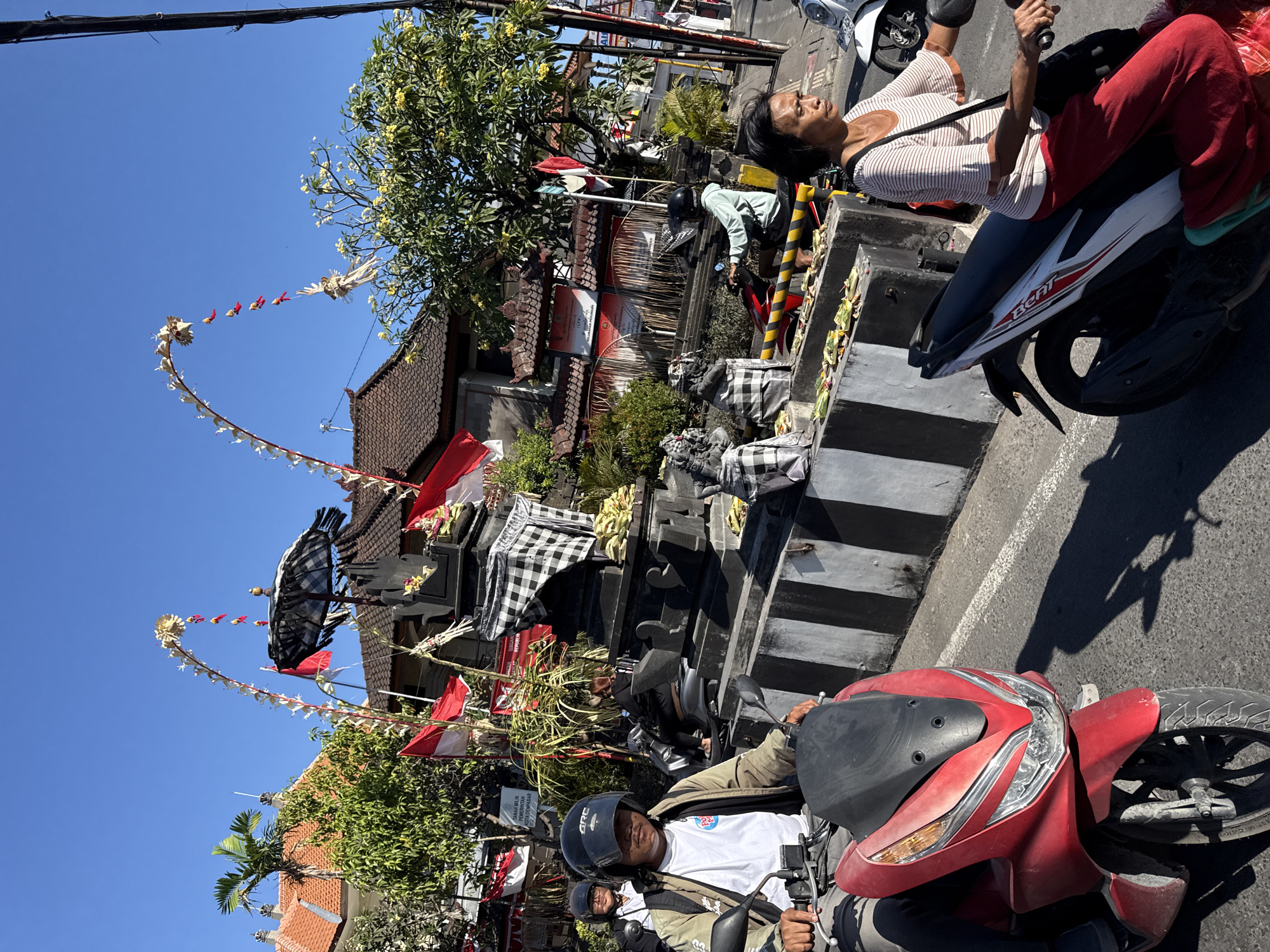 In Denpasar, Indonesia, on a sunny day, a man wearing a helmet and a beige jacket rides a red scooter. He is looking forward with a neutral expression. Behind him, another person on a scooter, also wearing a helmet, is visible. A black and white checkered umbrella is atop a stone monument, decorated with flags and offerings. The monument has a black and white striped base. Several people are on motorcycles and scooters, suggesting traffic. The environment includes buildings with red-tiled roofs, trees, and clear blue skies.