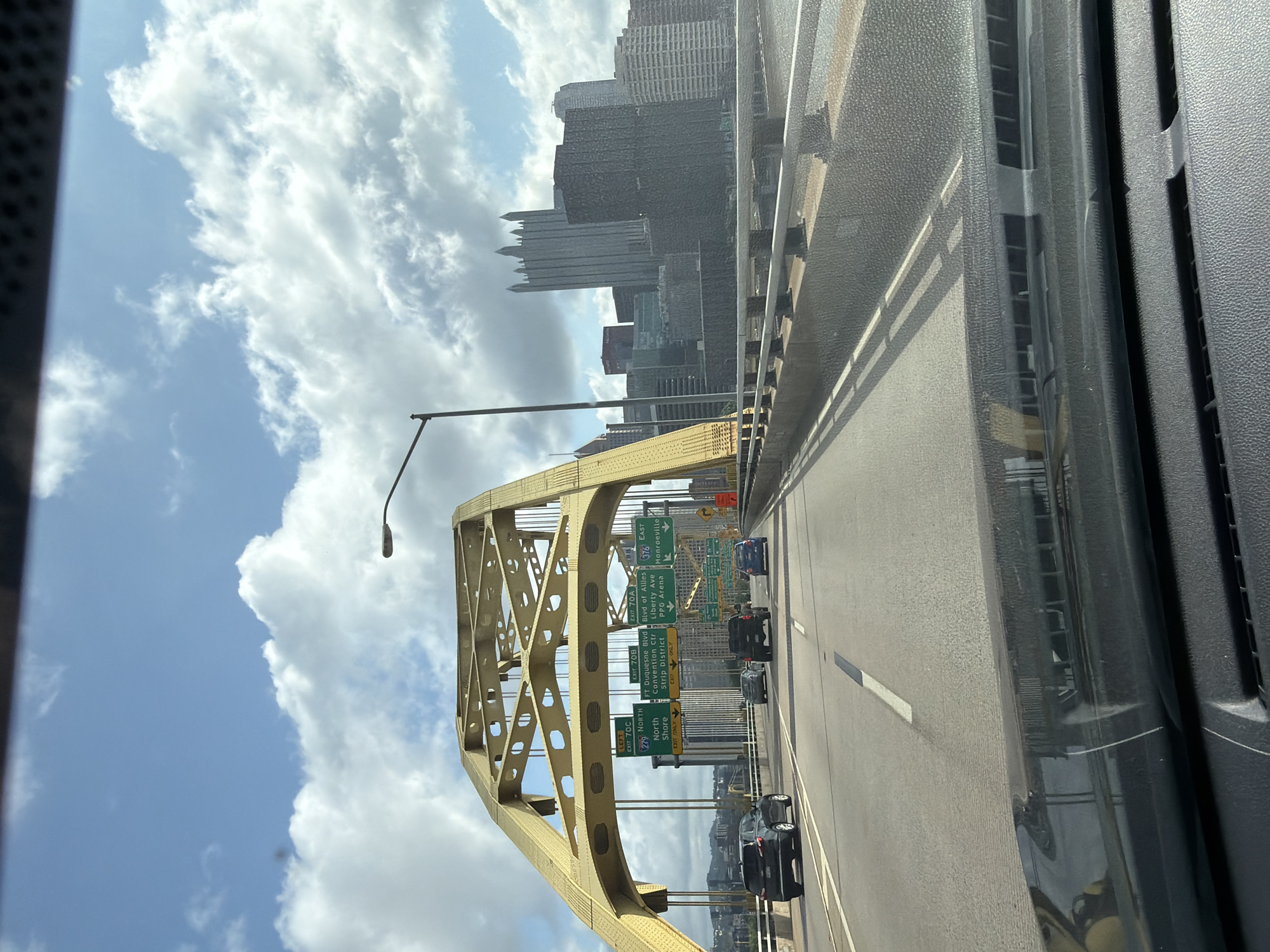 The image captures a daytime view from a vehicle driving on a bridge, likely a highway overpass, in Pittsburgh, Pennsylvania. A distinctive bright yellow suspension bridge arch dominates the upper portion of the frame, with its intricate steel latticework visible. Several highway signs are affixed to the bridge structure, indicating directions and exits: "LEFT EXIT 70C 276 NORTH North Shore", "FT. Duquesne Blvd Convention Ctr Strip District EXIT ONLY", "EXIT 70B", and "EAST 576 Liberty Ave PPG Arena Monroe ville". Beyond the bridge, the Pittsburgh skyline is visible, featuring several tall buildings, including a prominent gothic-styled skyscraper. The sky is partly cloudy with white cumulus clouds against a pale blue backdrop. Below the bridge, a multi-lane road carries traffic, with several cars seen on the highway. The foreground shows the interior of the vehicle, including the dashboard and windshield, reflecting the exterior scene. The environment suggests an urban setting with significant infrastructure.