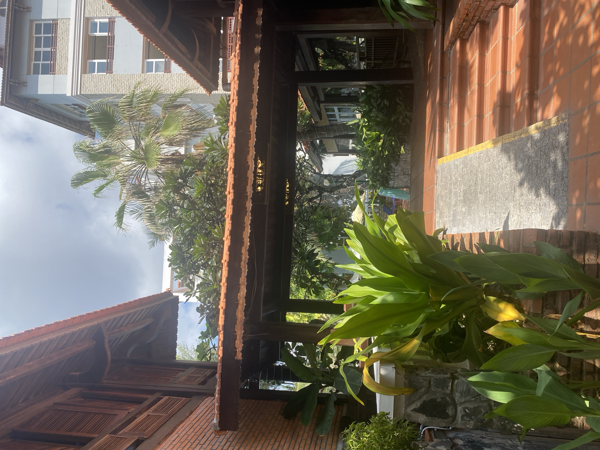 The image depicts an outdoor courtyard area within a resort or hotel complex in Phan Thiet, Vietnam. On the left, a traditional building with a brick base and dark wooden shutters and roof detailing is visible. To the right, a modern, multi-story building with light blue and stone facade and white framed windows stands prominently. A covered walkway with a red tile roof and dark wooden beams connects the two structures. Lush green tropical plants, including large-leafed plants and a palm tree, are abundant throughout the scene, suggesting a warm, sunny climate. The ground is paved with terracotta tiles, and a set of terracotta stairs leads up to the covered walkway. A grey concrete ramp with a yellow stripe runs alongside the stairs. The sky is partially cloudy, with bright sunlight illuminating the scene. No people are visible in the image.