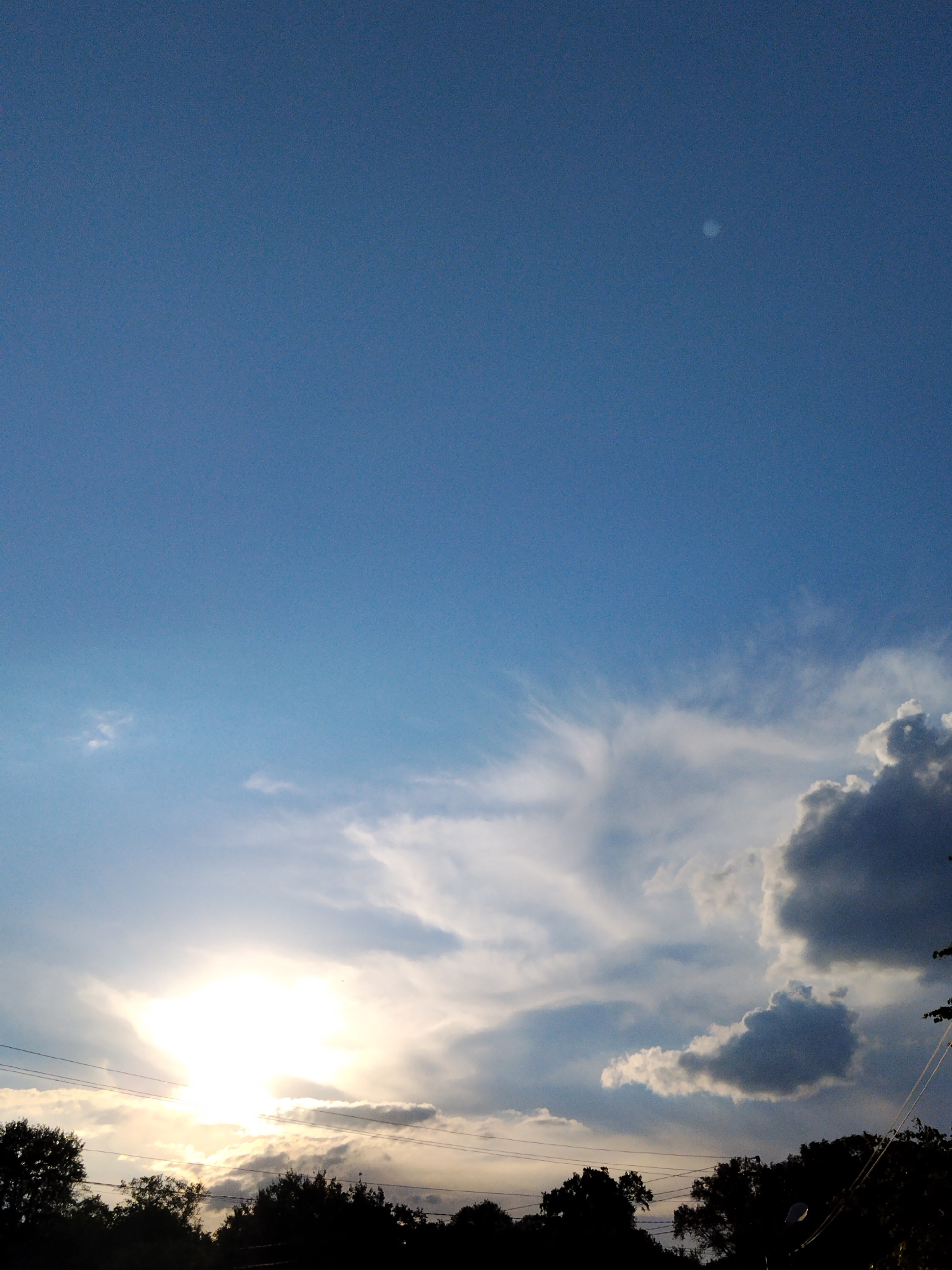 This is a vertical, outdoor, medium-shot photograph of a sky during what appears to be either sunrise or sunset. The upper portion of the image is dominated by a clear blue sky, with a small, pale, round object, possibly the moon or a distant planet, visible in the top right quadrant.
As the eye moves down, the sky transitions to include various cloud formations. On the left, a bright, blown-out area indicates the sun, partially obscured by wispy white clouds and some darker, heavier clouds. The sunlight casts a warm glow, suggesting the golden hour. In the lower half of the frame, silhouetted trees form a dark band across the bottom, creating a stark contrast with the bright sky. Power lines are also visible stretching horizontally across the lower part of the image, in front of the trees and the sun.
The weather appears partly cloudy, with a mix of dramatic dark clouds and softer, luminous white clouds. The presence of the sun low in the sky, combined with the silhouetted trees, strongly suggests either early morning or late evening. There are no people or specific objects, other than the natural elements of the sky, trees, and utility lines, visible in the frame. No text is present in the image. The overall mood conveyed is one of natural beauty and the dramatic transition of light.