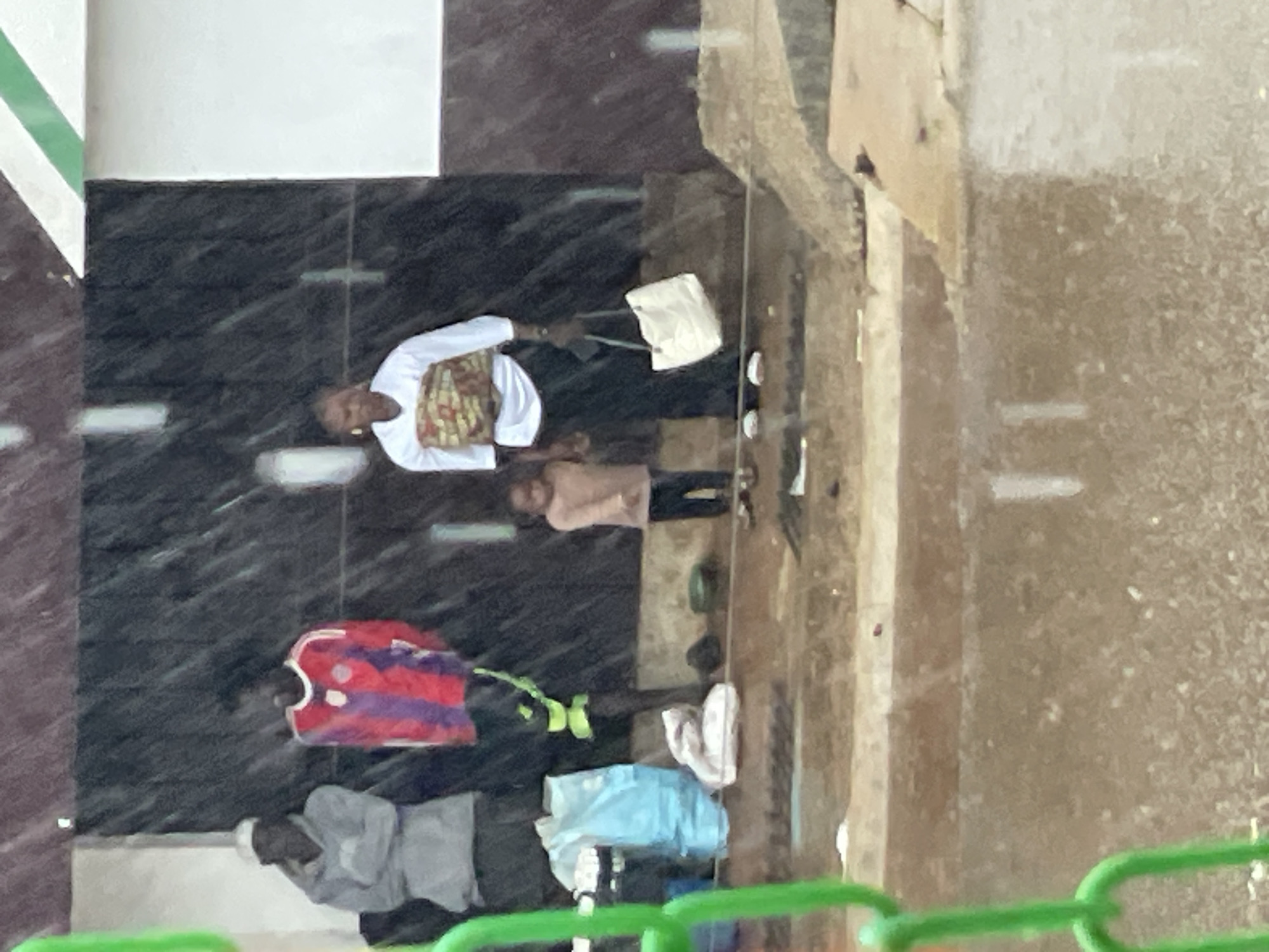 In the city of Unknown, Unknown, under rainy conditions, several individuals are gathered near a dark, tiled structure with some green and white accents at the top. In the foreground, a person wearing a grey hoodie and a white cap stands with their arms crossed. To their right, another person is wearing a red and purple striped long-sleeved shirt. A young child in a pink long-sleeved shirt and dark pants stands next to an adult woman wearing a white long-sleeved shirt and a patterned garment around her waist. The woman is holding a white tote bag. In the background, there are some bags and other objects on the ground. The ground appears wet and muddy, suggesting recent rain or standing water. Green fencing is visible in the extreme foreground, partially obscuring the scene.