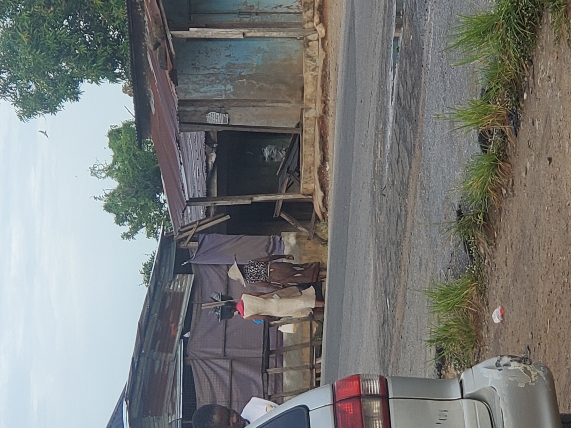 The image shows a street scene in an unidentified city. On the left, there is a dilapidated structure with a corrugated iron roof and fabric covering the walls. Two women are standing near the structure, with one wearing a straw hat and a dress. A silver car is visible in the foreground. The street is paved and has grass growing along the edge. A person can be seen partially visible in the very bottom of the frame, next to the car.
