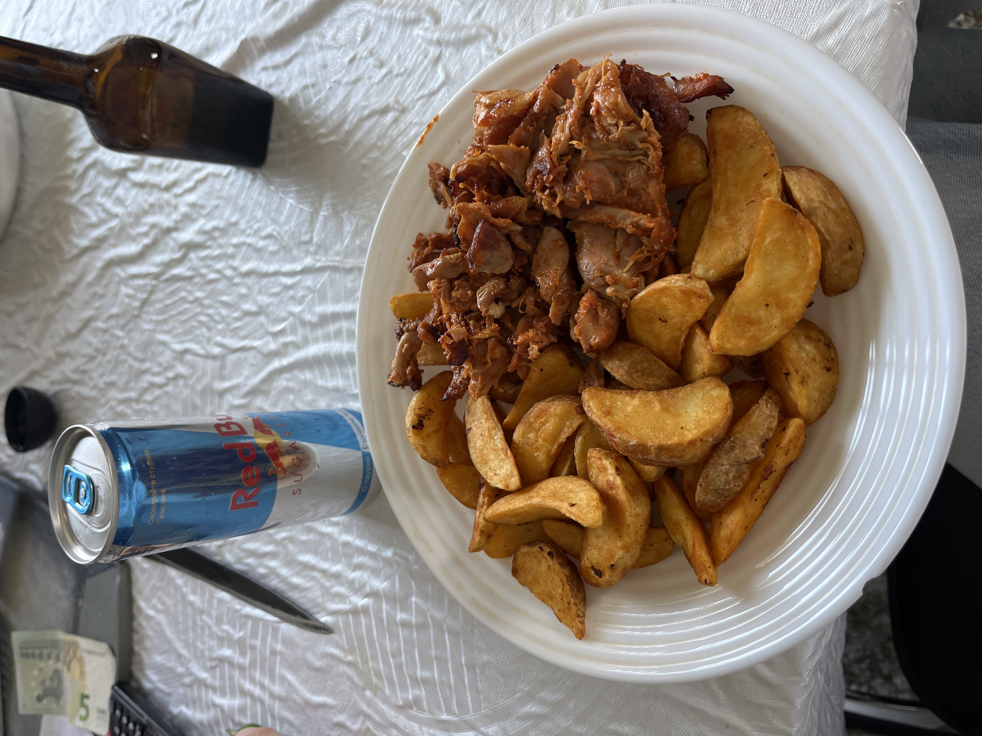 An overhead shot captures a meal laid out on a white, textured tablecloth. The central focus is a large white plate filled with food. On the left side of the plate is a generous portion of cooked meat, appearing shredded or chopped, with a reddish-brown, slightly crispy texture, resembling gyros or döner kebab. To the right of the meat are numerous golden-brown potato wedges, some with visible seasoning.
To the left of the plate, a silver and blue Red Bull Sugarfree can is lying horizontally. Visible text on the can includes "Red Bull" and "SUGARFREE" in English, along with Cyrillic text that reads "СЪБУЖДА ВАШИТЕ СЕТИВА" (Awakens your senses), "Газирана напитка" (Carbonated drink), and "със таурин и кофеин" (with taurine and caffeine). Further to the top-left, a dark amber glass bottle, possibly a beer or spirit bottle, lies partially visible.
In the bottom-left corner, a fragment of a Bulgarian 5 lev banknote is visible, showing the numeral "5" and part of the design. A dark object, possibly a remote control or electronic device, is also partially seen. The scene suggests a casual meal setting, likely indoors, consistent with the provided location context of Zlatograd, Bulgaria, given the visible Cyrillic text and currency. The overall lighting suggests daylight, but the exact time of day is not discernible.