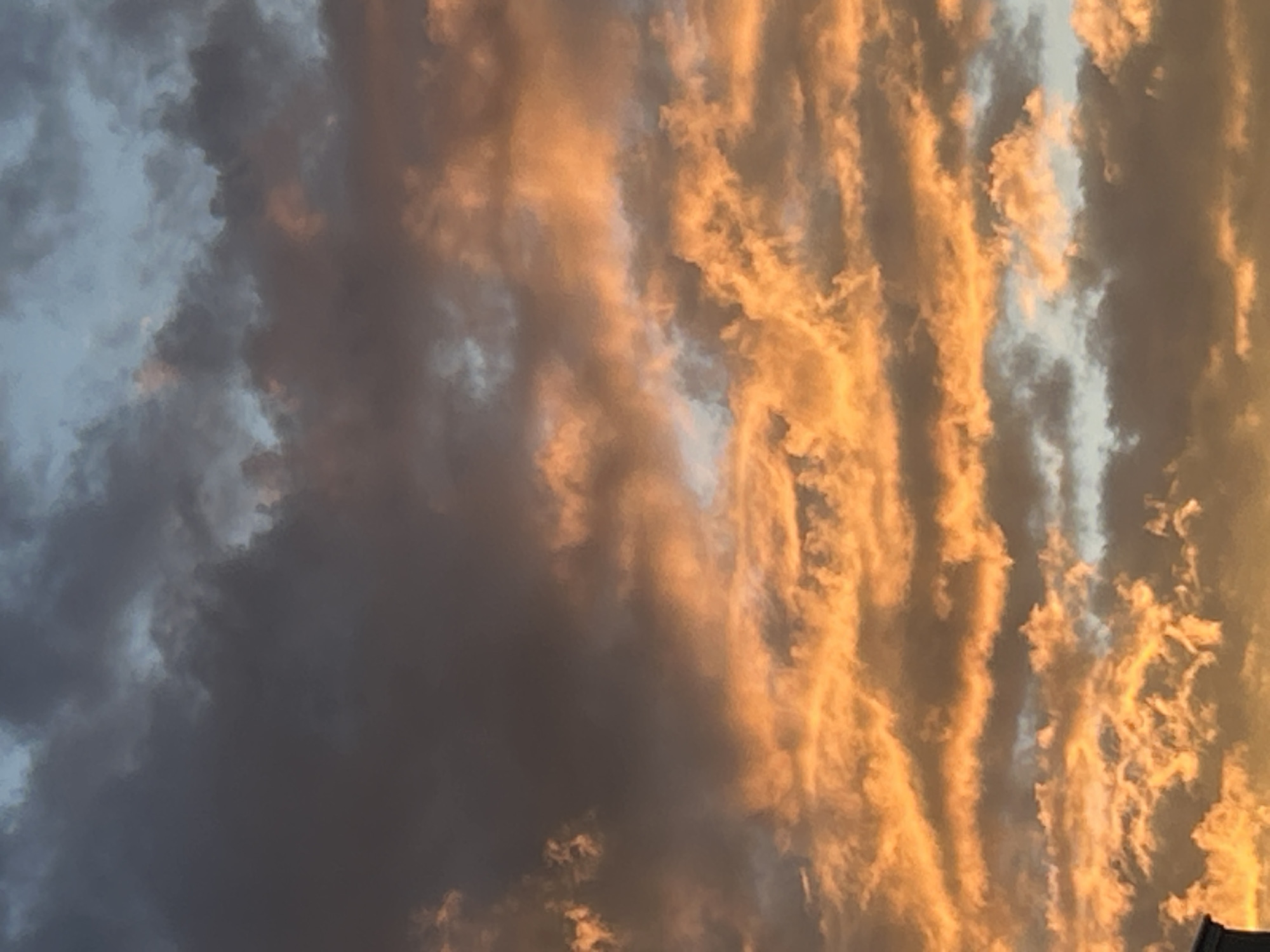 The image depicts a sky filled with dramatic clouds illuminated by the warm hues of a sunset. The clouds are a mix of dark grays and vibrant oranges, creating a striking contrast. Some clouds are thin and wispy, while others are dense and voluminous. The light suggests it is either sunrise or sunset. A dark structure, likely a building, is partially visible at the bottom left corner of the image, suggesting an urban setting. The scene is devoid of people or any discernible activity. The image does not provide any specific visual cues that would allow for identification of the location within the city of Plovdiv, Bulgaria.