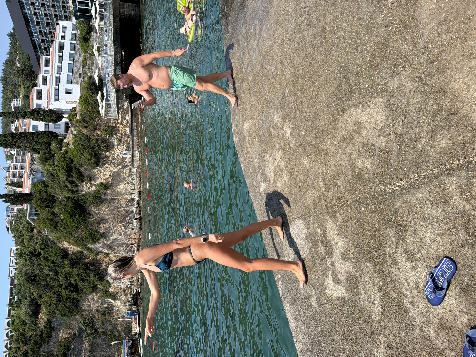 In Budva, Montenegro, on a sunny day, a young woman in a dark teal bikini walks along a concrete pier, pointing to her left with her left hand. She has blonde hair and is wearing a watch on her left wrist. Behind her, a man in green swim trunks stands on the pier, looking at his phone. He is shirtless and has short brown hair. In the background, the turquoise water of the Adriatic Sea is filled with people swimming and relaxing on inflatable floats. Buildings with white facades and red roofs are visible on a lush green hillside overlooking the water, with a modern hotel structure on the far right. The pier is textured concrete, with a pair of blue flip-flops lying on it in the foreground.