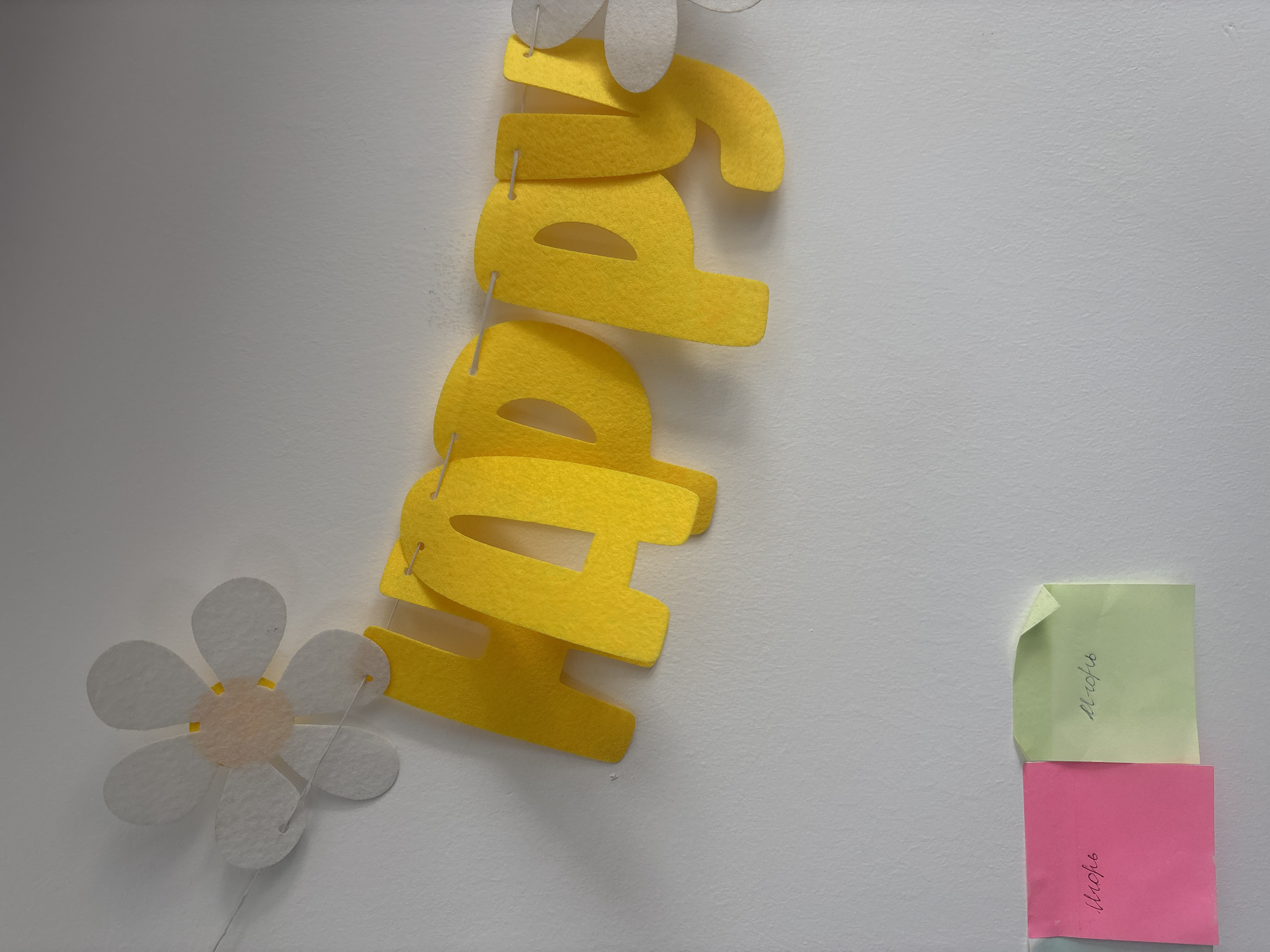 In a close-up, eye-level shot, a white wall serves as the backdrop for a "Happy" banner made of yellow felt letters strung together with white twine. The banner is positioned diagonally across the upper portion of the frame. To the left of the banner, a white felt flower decoration with a light pink center is attached to the wall. To the right, another white felt flower decoration is partially visible. Below the banner, two sticky notes are affixed to the wall. The left note is pink and has handwritten text in cursive that appears to be "Школы". The right note is light green and has handwritten text in cursive that appears to be "Школы". The lighting is consistent and the image is in focus. The scene depicts a decorated wall, possibly in a classroom or a child's room.