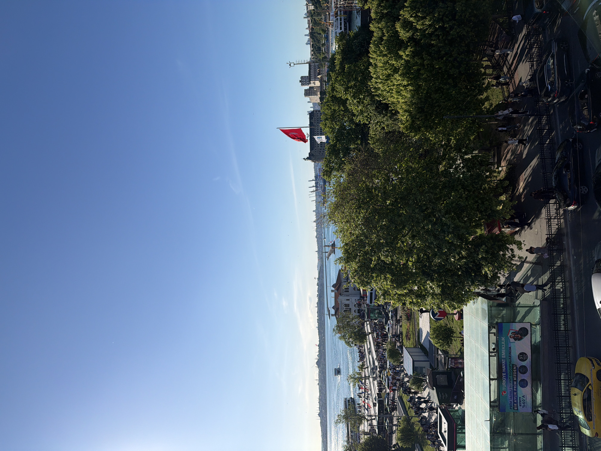 The image shows a cityscape with a waterfront view, likely taken from an elevated position. The scene includes a blue sky, a body of water (likely the Bosphorus), and buildings of varying heights in the distance. A Turkish flag is prominently displayed.
In the foreground, there's a busy street with trees, cars, people walking, and what appears to be a bus stop. There's also a yellow taxi visible.
Given the Turkish flag and the cityscape characteristics, the location is very likely within Istanbul, Turkey. The features match the Kadıköy district on the Asian side, with its views of the Bosphorus and active city life.