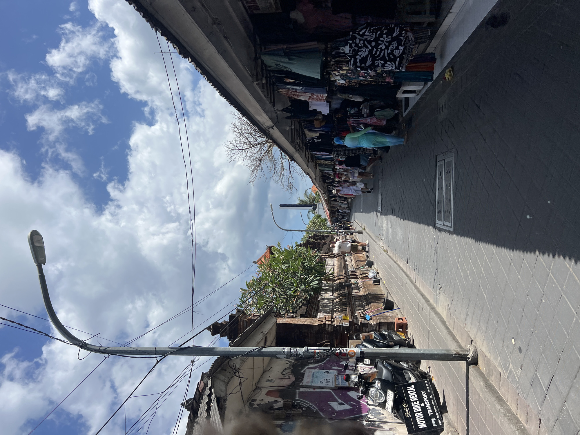 This is a street scene in Kuta, Indonesia, on a bright, cloudy day. The street is paved with gray interlocking bricks. On the left, a building with a graffiti-covered wall and an ornate brick facade is visible, with a parked black motorcycle with a "Bali Car Motor Bike Rental & Transport" sign in front of it. A street lamp with a curved arm rises from the left side of the image, with a tangle of electrical wires overhead. Further down the street on the left, a few trees with green foliage are planted along the wall, and some people are walking. On the right side of the street, a row of open-air market stalls displays clothing for sale, with racks of garments hanging under a corrugated roof. Several people are browsing the merchandise and walking along the street. The sky is blue with scattered white clouds.