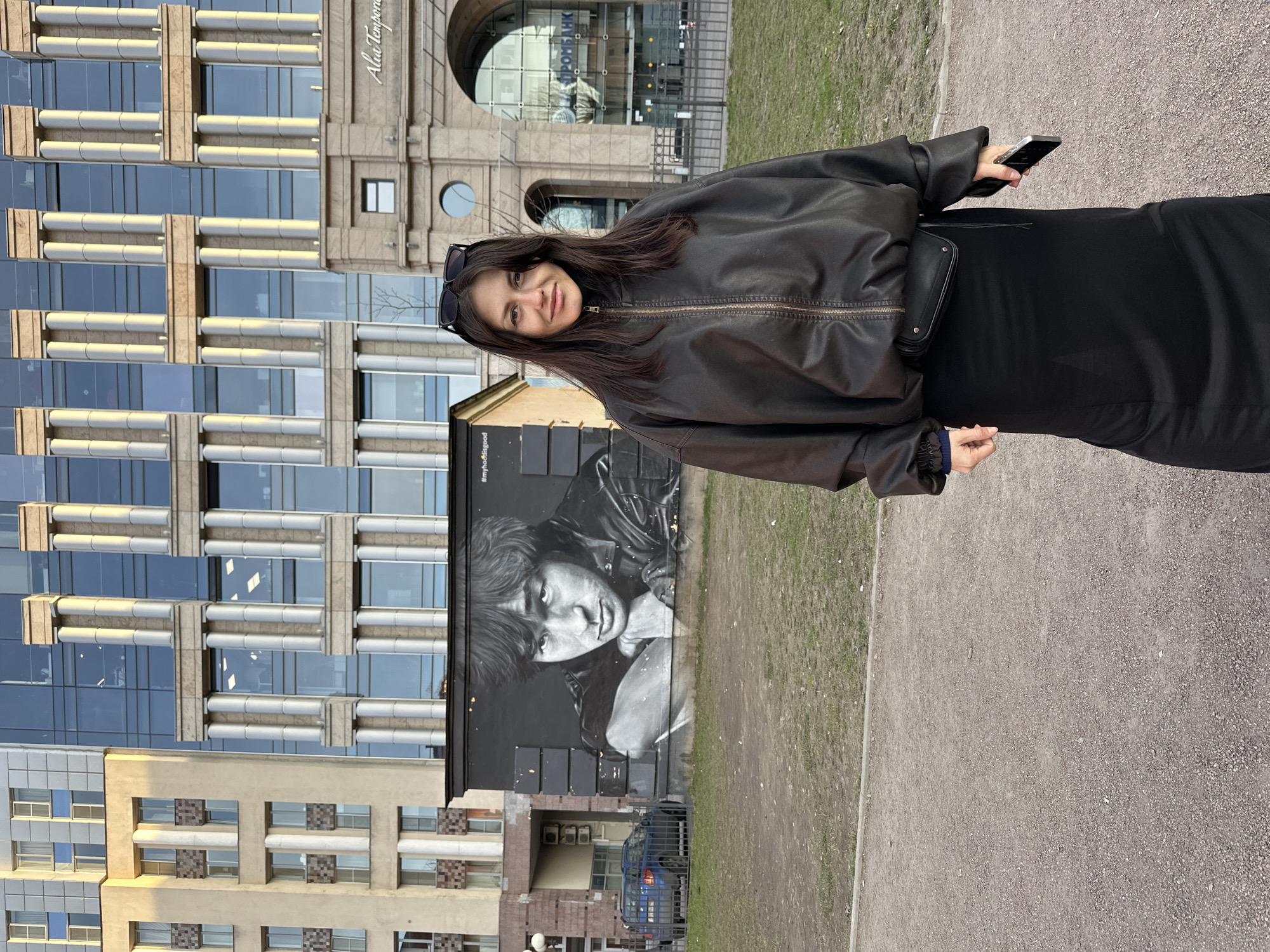 An outdoor, eye-level full shot captures a young woman standing on a gravel path with urban architecture in the background. The woman, with long dark hair and sunglasses perched on her head, wears a black leather jacket and a dark dress. She looks directly at the camera with a subtle smile, holding a silver-cased smartphone in her right hand.
Behind her, to the left, a prominent black and white mural depicts a close-up portrait of a young man, possibly in a leather jacket, with a serious expression. Above the mural, the hashtag "#myhoolieisgood" is visible. The mural is painted on a building with a simpler, possibly older, facade.
Further to the left, a modern, multi-story building rises with a striking glass facade interspersed with decorative light-colored vertical and horizontal elements. Partial text, "Aura Tempora," can be seen on this building. To the right of the woman, in the mid-ground, stands a more traditionally designed building with an arched entrance and classical architectural details. The name "ПРОБАНК" (PROBANK) is clearly visible on its signage.
The ground consists of a light-colored gravel path bordered by dry, patchy grass. The scene is illuminated by natural daylight, suggesting clear weather. The setting is an urban environment, specifically Saint Petersburg, Russia.