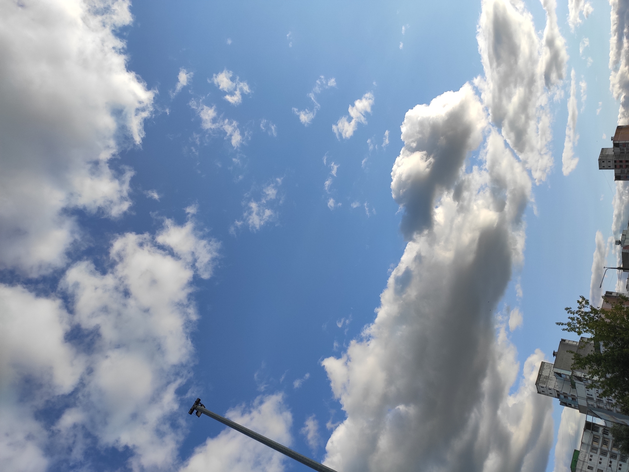 A wide-angle, outdoor, medium shot captures a sky dominated by blue with scattered white and grey clouds. A tall, slender, grey metal pole with a fixture at the top extends diagonally from the left side of the frame into the upper sky. In the lower section of the image, the tops of several multi-story buildings are visible, along with some green foliage. The architecture suggests an urban environment. The scene is consistent with a view from the city of Homyel, Belarus, likely in a residential or mixed-use area. The lighting indicates daytime, with the sun illuminating the clouds and casting shadows on the buildings. No people or actions are depicted.