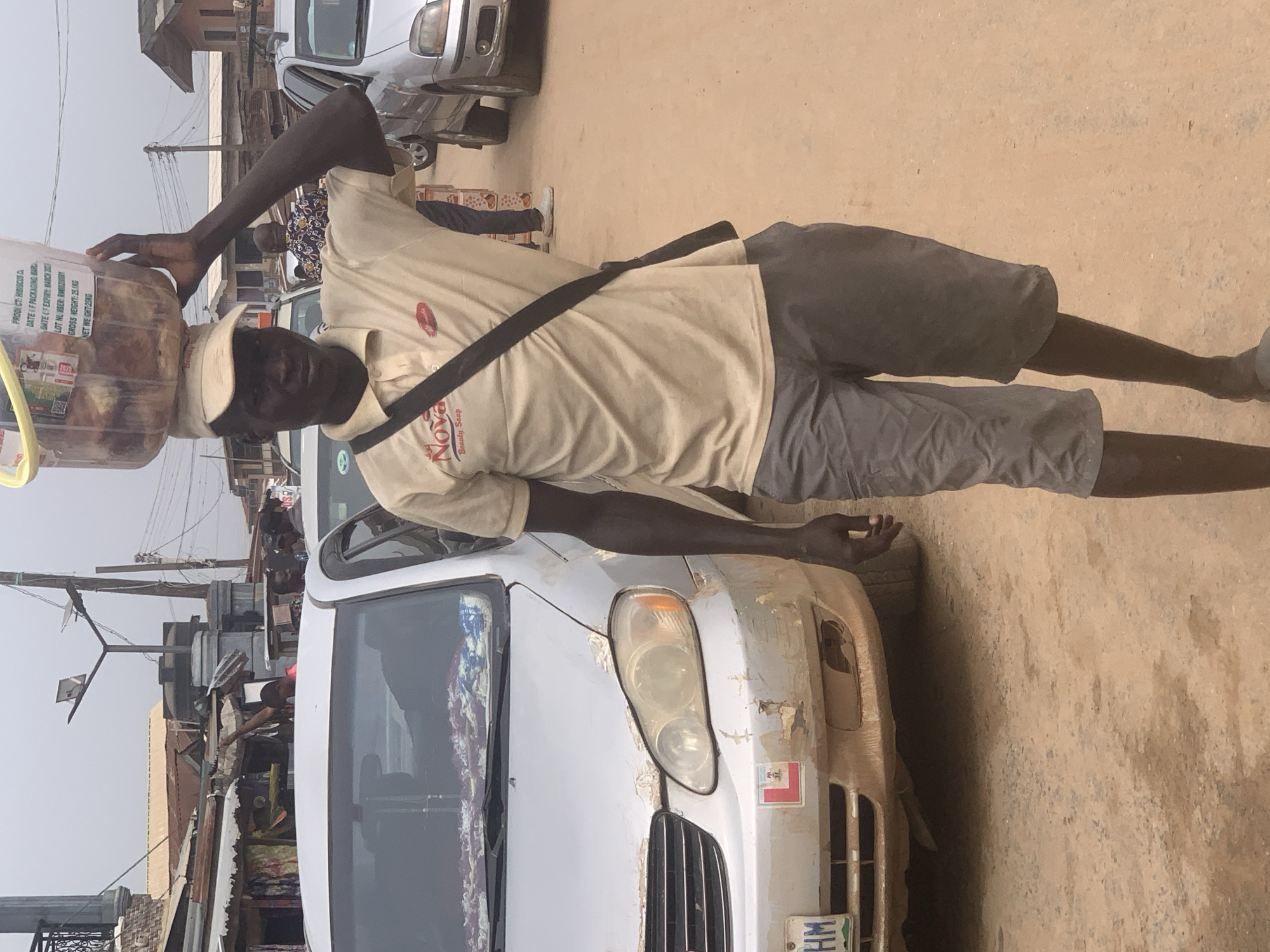 The image depicts a man in Shagamu, Nigeria, standing in front of a silver car with damage to the front bumper and passenger side headlight. The man is wearing a tan polo shirt with the logo "Nova Beauty Soap", gray shorts, and a cap. A black strap goes across his body from his left shoulder to his right hip. He balances a clear plastic container on his head. The container is labeled "PRODUCT: HIBISCUS C," with a date of packaging and an expiry date of March 2025, and a gross and net weight of 25.1kg and 25kg, respectively. The environment appears to be a busy street with other vehicles and buildings visible in the background. The ground is dirt. Electrical poles and wires are overhead.