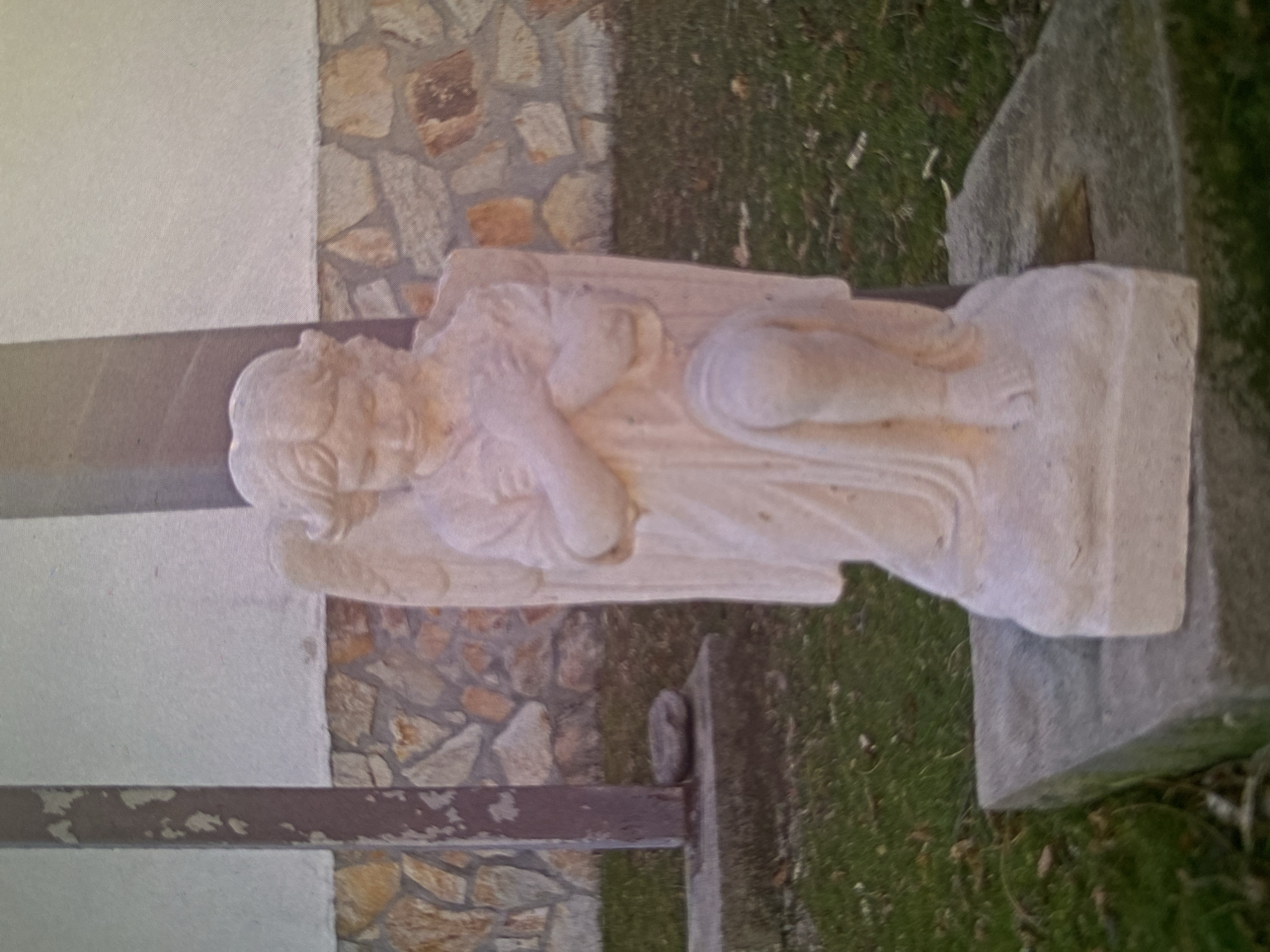 The image features a light-colored stone statue of a serene angel, situated outdoors in Bankya, Bulgaria. The angel is depicted lying down on its side, appearing to rest peacefully. Its head is gently supported by its left arm, and its right arm is crossed over its chest. The angel's face shows a calm expression with closed or downcast eyes, and sculpted hair frames its face. Detailed wings are visible on its back, and flowing robes are draped around its body, with legs slightly bent.
The statue rests on a rectangular, light-colored stone base, which is positioned on the right side of the frame. The background consists of architectural elements and natural ground cover. To the left of the statue, there is a plain, light-colored wall. Adjacent to this and continuing behind the statue is a wall section adorned with irregular stone cladding in various shades of brown and grey. Below the statue and extending to the right, areas of green grass are visible, suggesting an outdoor garden or yard setting. A dark, possibly metal, bar or railing runs horizontally across the lower left part of the image, partially obscured.
The scene is illuminated by natural daylight, indicating it is daytime, with soft lighting evident from the lack of harsh shadows. No text is visible in the image. The overall atmosphere conveyed is one of peacefulness and contemplation, with the statue serving as a focal point within its natural and constructed surroundings.