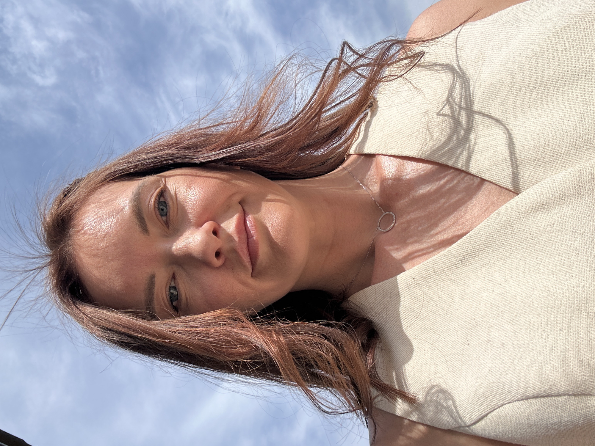 A photograph depicts a woman from a low angle, seemingly looking downward at the camera. The background is a bright blue sky scattered with cirrus clouds. The woman has fair skin, auburn hair, and green eyes. She is wearing a light beige sleeveless top and a simple silver necklace with a circular pendant. The light is bright, causing the skin to appear somewhat reflective and emphasizing the texture of the clothing. The location appears to be outdoors, but there is no other indication of where it is located other than that the user has requested that it be identified as Прикамский, Russia.