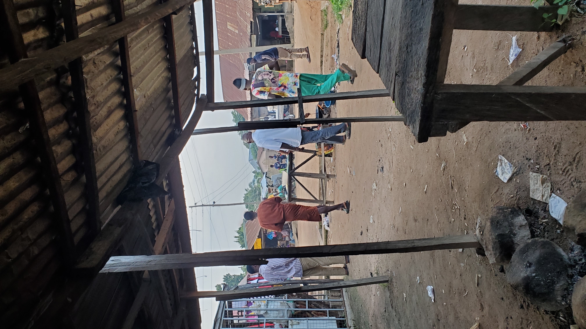 This image depicts a street scene in Jalingo, Nigeria, under a rudimentary shelter with a corrugated metal roof supported by wooden beams. In the foreground, a rustic wooden table is positioned. The ground is unpaved and appears dusty, with scattered debris including paper and plastic.
Several individuals are visible in the scene. On the left, two men stand near a stall with shelves displaying various items, one wearing a striped shirt and beige trousers, the other in an orange garment. Further into the scene, a man in a white shirt and grey trousers walks away from the camera. To the right, a man in a colorful shirt with a pattern and green trousers walks with his back to the camera, and another man in a dark shirt stands further back. In the middle ground, other individuals are seen sitting and standing near wooden structures and what appear to be shops. The background shows more buildings with corrugated metal roofs and some vegetation, suggesting a semi-urban or rural environment. Power lines are also visible overhead. The overall impression is of a bustling outdoor market or community gathering area.