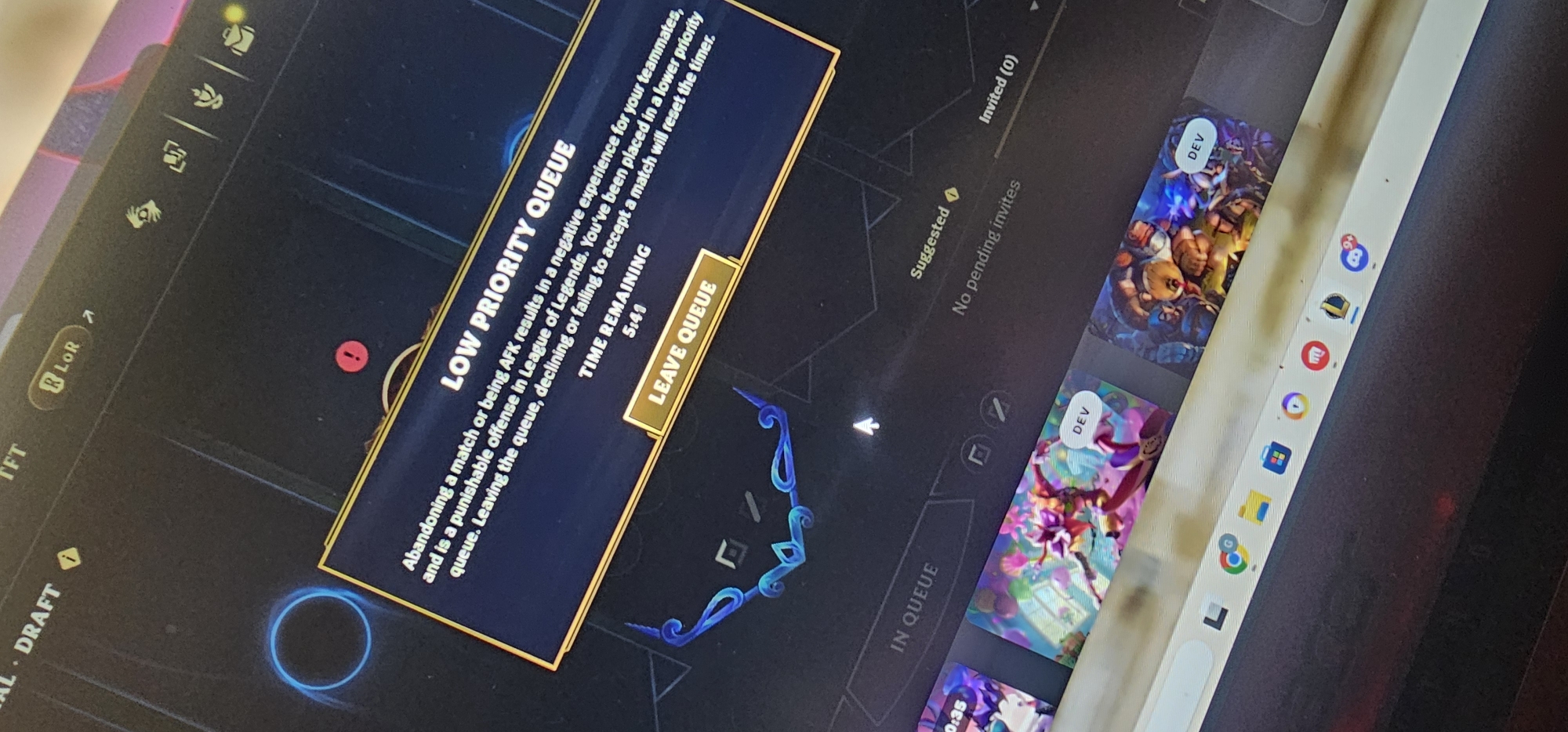 The image shows a computer screen displaying the "Low Priority Queue" message from the game League of Legends. The message explains that abandoning matches or being AFK results in a negative experience for teammates and is a punishable offense. The player is placed in a lower priority queue, and leaving the queue, declining, or failing to accept a match will reset the timer. The remaining time is 5:41. The "Leave Queue" button is visible. The screen also shows elements of the game's interface, including queue status, suggested friends, and pending invites. Icons for other applications such as Chrome, Microsoft Edge, and Riot Vanguard are visible on the taskbar. There are also some small specks of dirt on the screen near the taskbar. The photo was taken at an angle, and the location is not provided in the image.