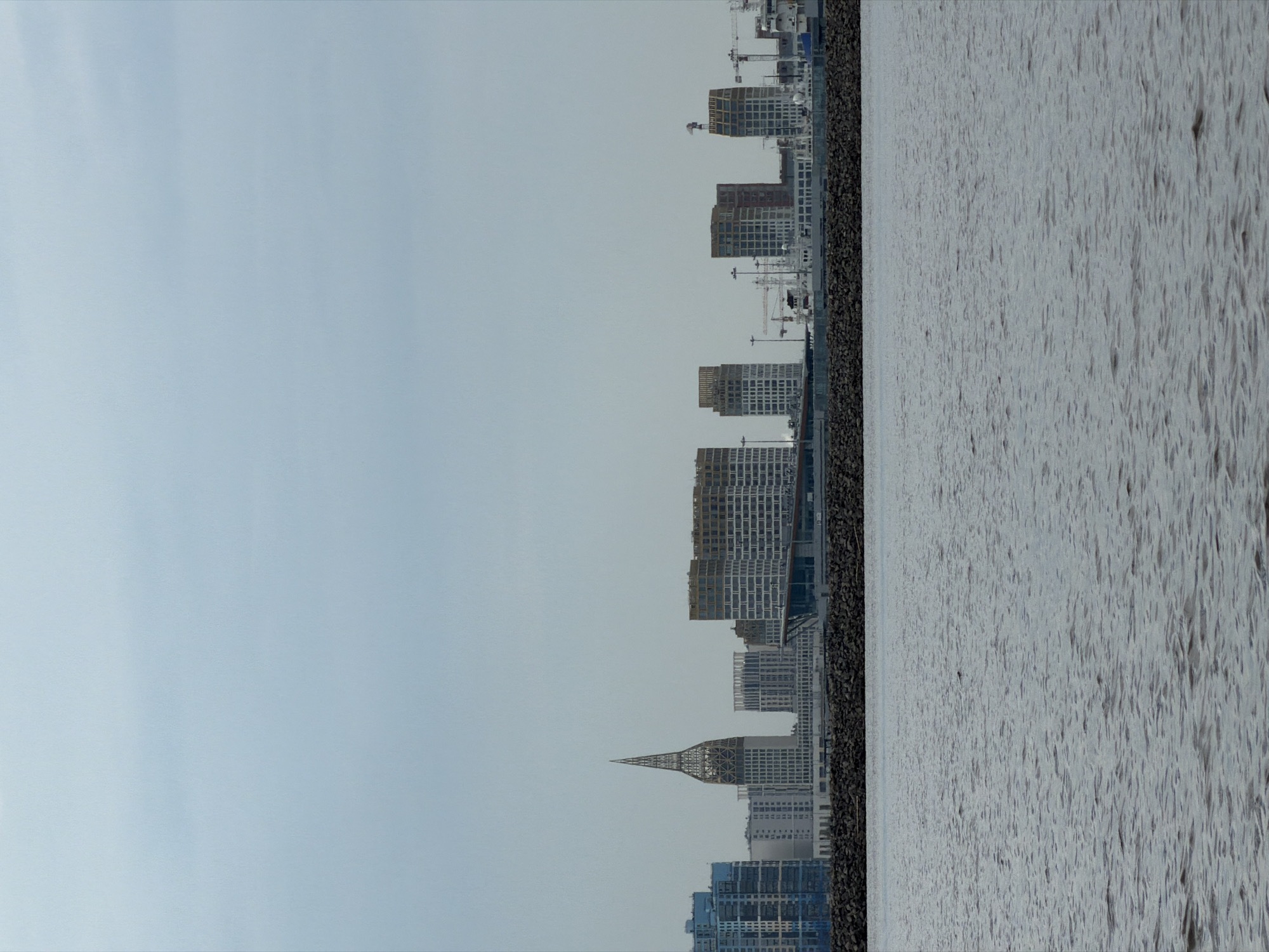 The image shows a cityscape with tall buildings and cranes, suggesting ongoing construction. The buildings are modern, with varied architectural styles, including some with repetitive window patterns and others with more unique designs, such as a spire-like structure. The scene is set against a pale, overcast sky. To the right of the buildings is a body of water, likely a river or bay, with a textured surface, indicating gentle waves or ripples. A dark, textured barrier, possibly a retaining wall or embankment, separates the buildings from the water. The overall impression is one of urban development and a waterfront environment. The lighting suggests it is daytime, possibly morning or late afternoon, given the diffused light. There is no visible text in the image.