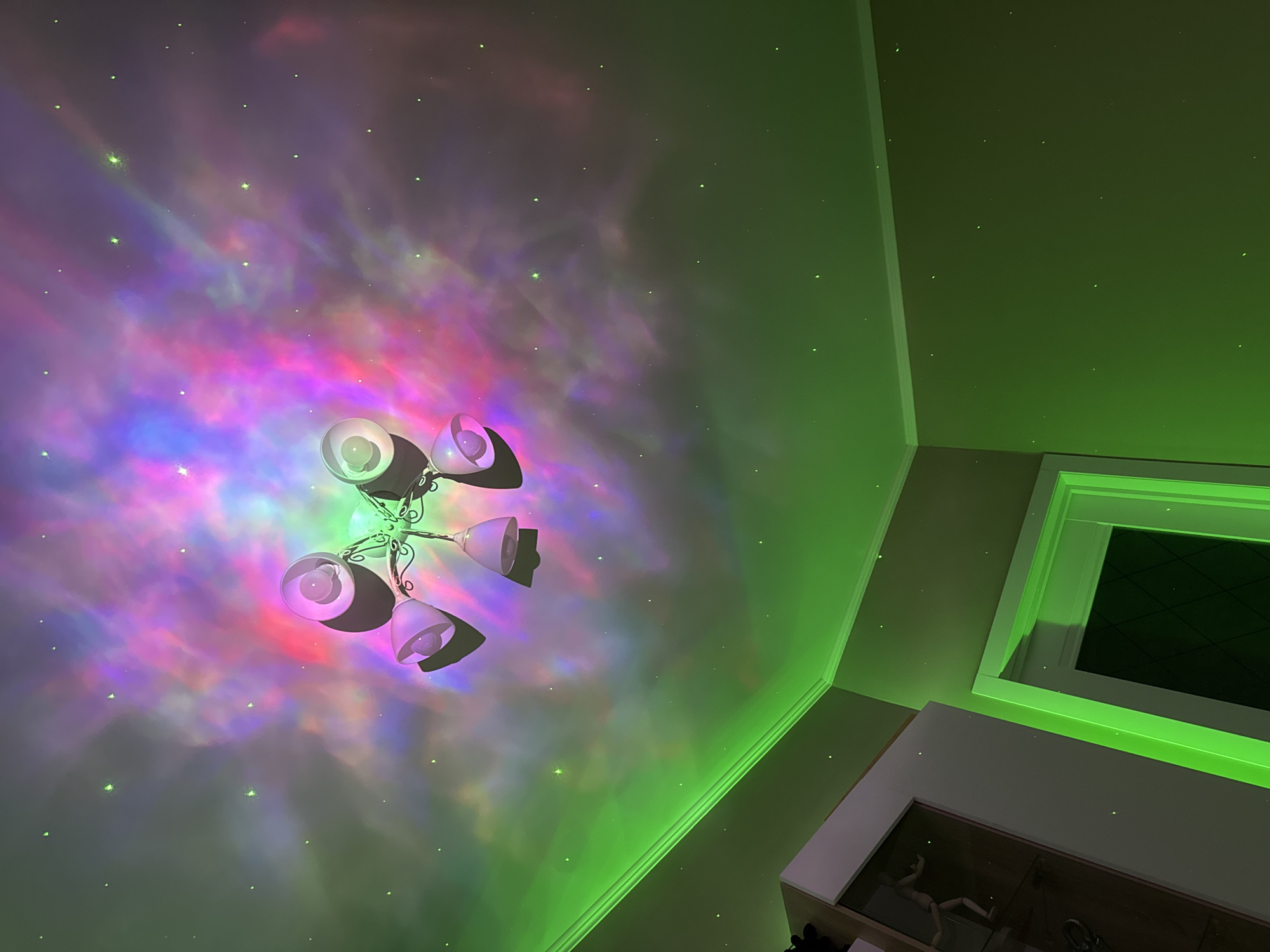 The image captures an interior scene under artificial lighting. A decorative light fixture with multiple lamps is positioned on the ceiling. The light projects a nebula-like pattern with hues of purple, red, and blue across the ceiling. The walls are illuminated with a strong green light. A section of the wall with a window is visible, also lit with green light. There are small, bright dots scattered across the illuminated surfaces, resembling stars. The location is Unknown, Unknown.